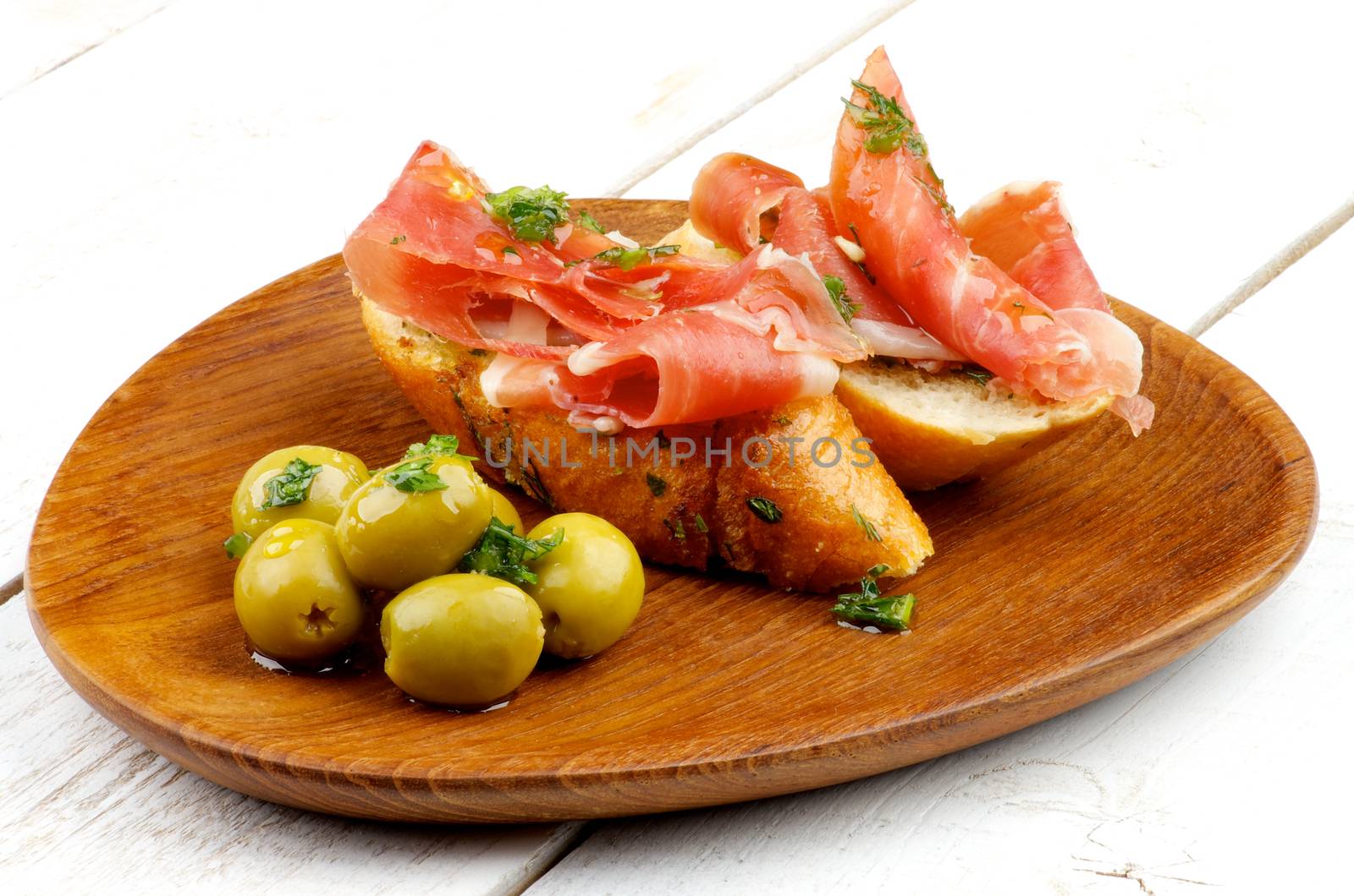 Delicious Tapas with Smoked Jamon, Garlic Bread and Green Olives on Wooden Plate closeup on White Wooden background