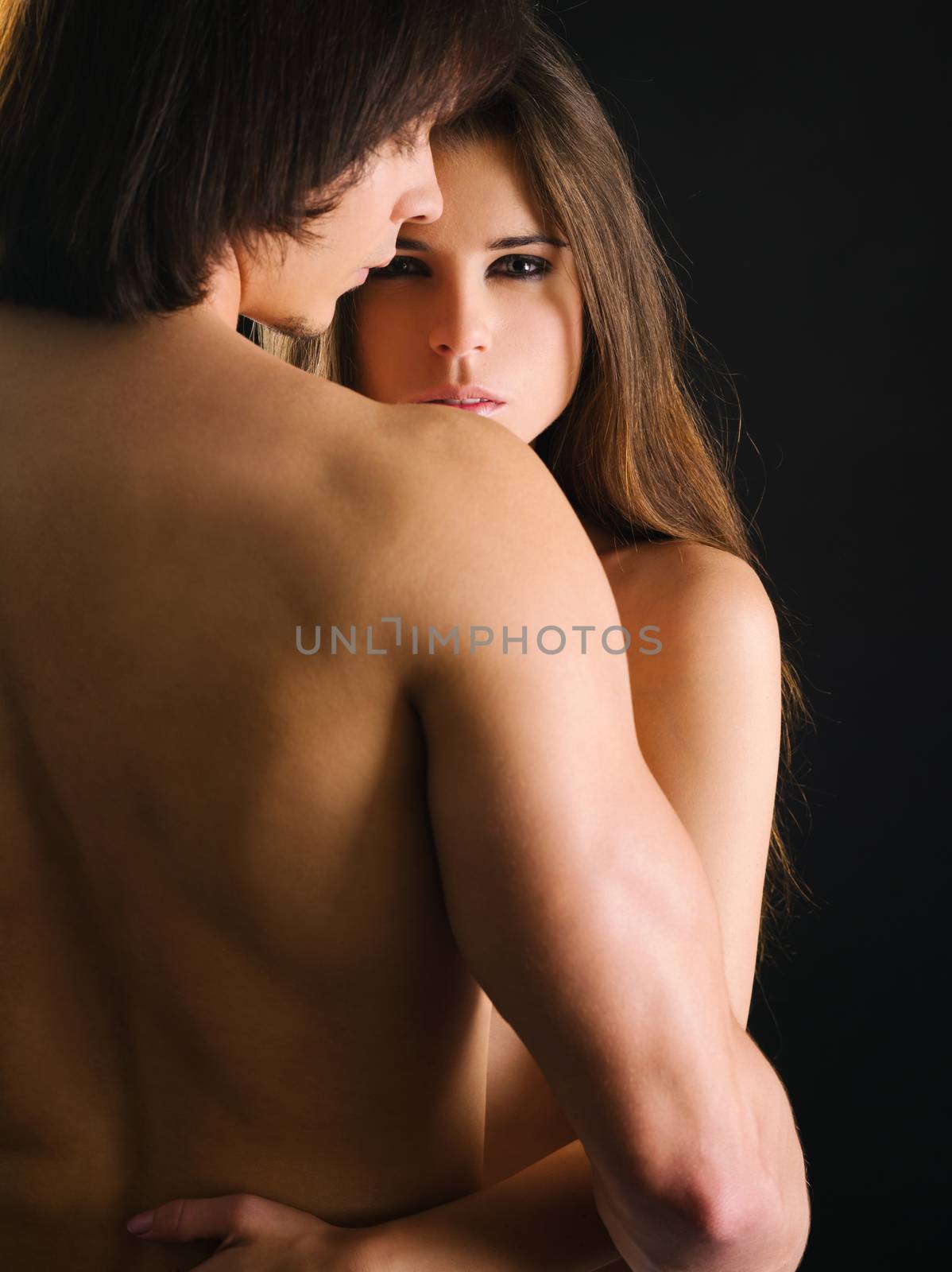 Photo of a topless young woman and man in love and holding each other. Woman is staring at the camera.