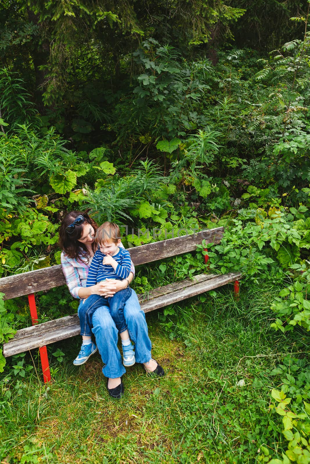 Photo of a young mother and her son sitting on a park bench surrounded by trees and bushes.