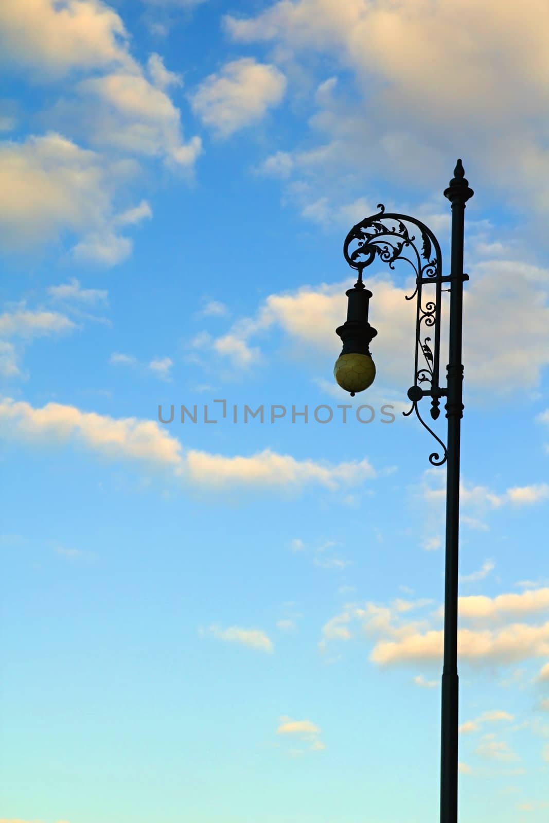 Photo shows detail of street lamp in front of the white clouds and blue sky.