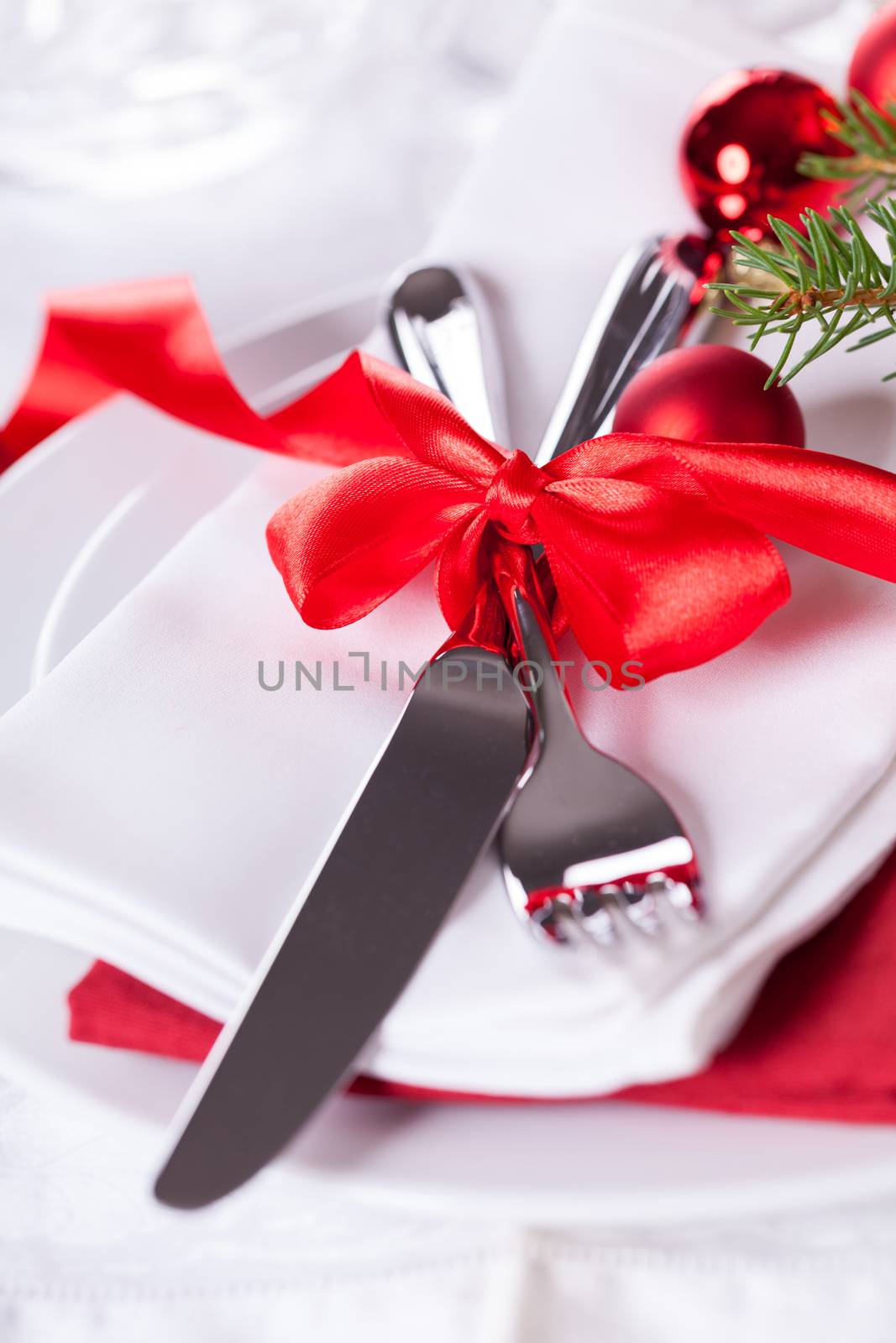 Romantic red Christmas table setting with white plates, red and white linen and silverware tied with a red ribbon and bow decorated with red Xmas baubles and evergreen natural pine foliage