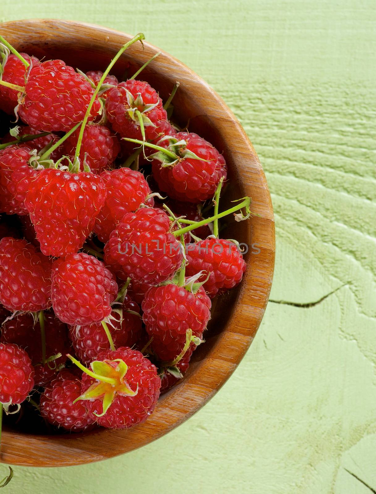 Perfect Ripe Raspberries with Stems in Wooden Bowl on Light Green Wooden background. Top View