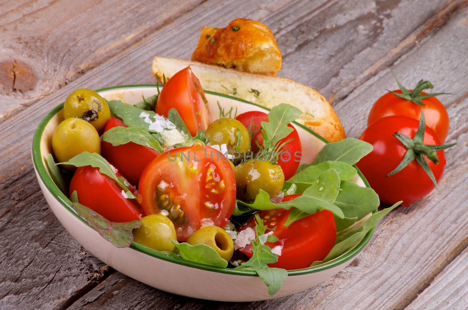 Arrangement of Fresh Tomatoes Salad with Arugula, Olives, Greens and Garlic Bread isolated on Rustic Wooden background