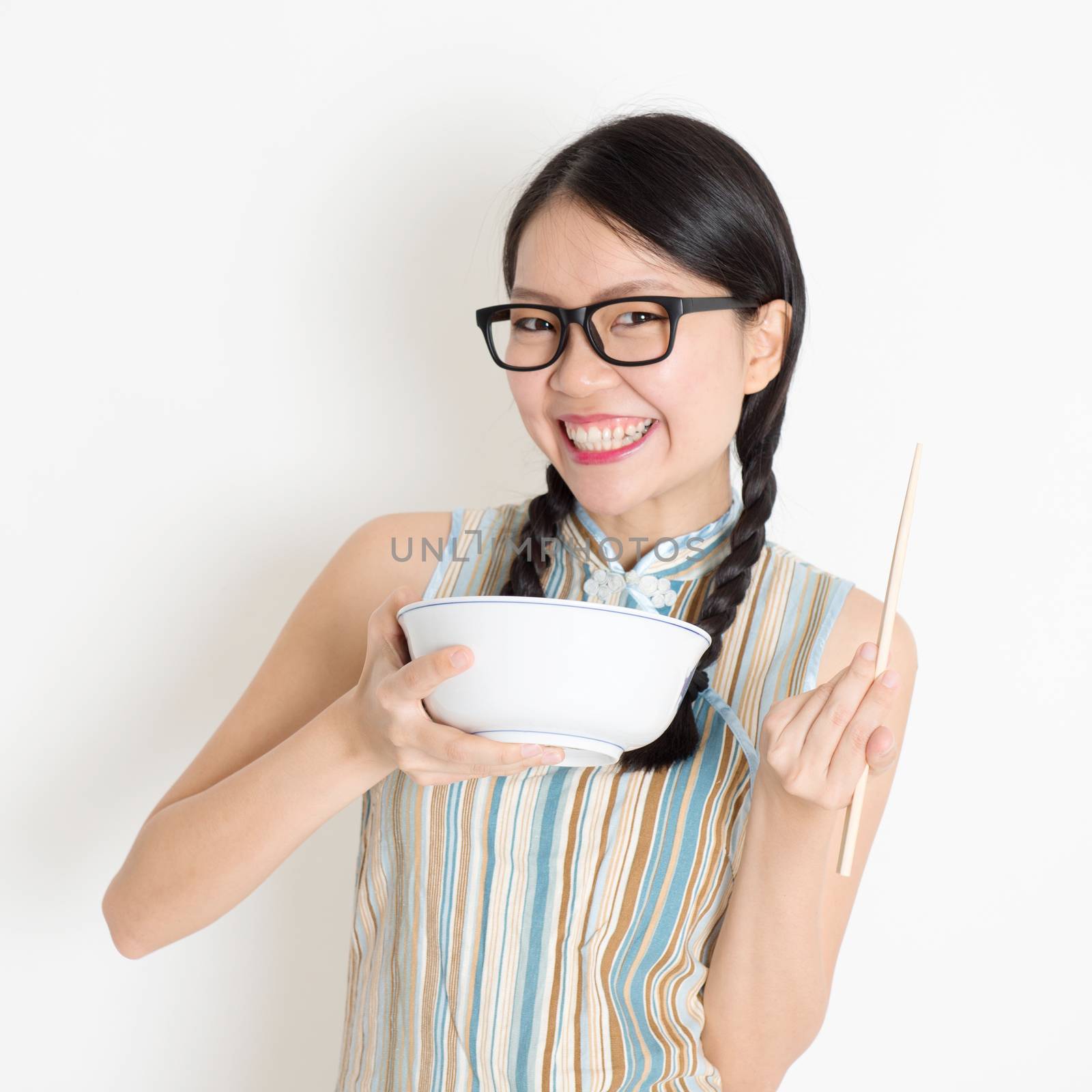 Portrait of Asian Chinese girl eating, using chopsticks holding rice bowl, in retro revival style cheongsam standing on plain background.