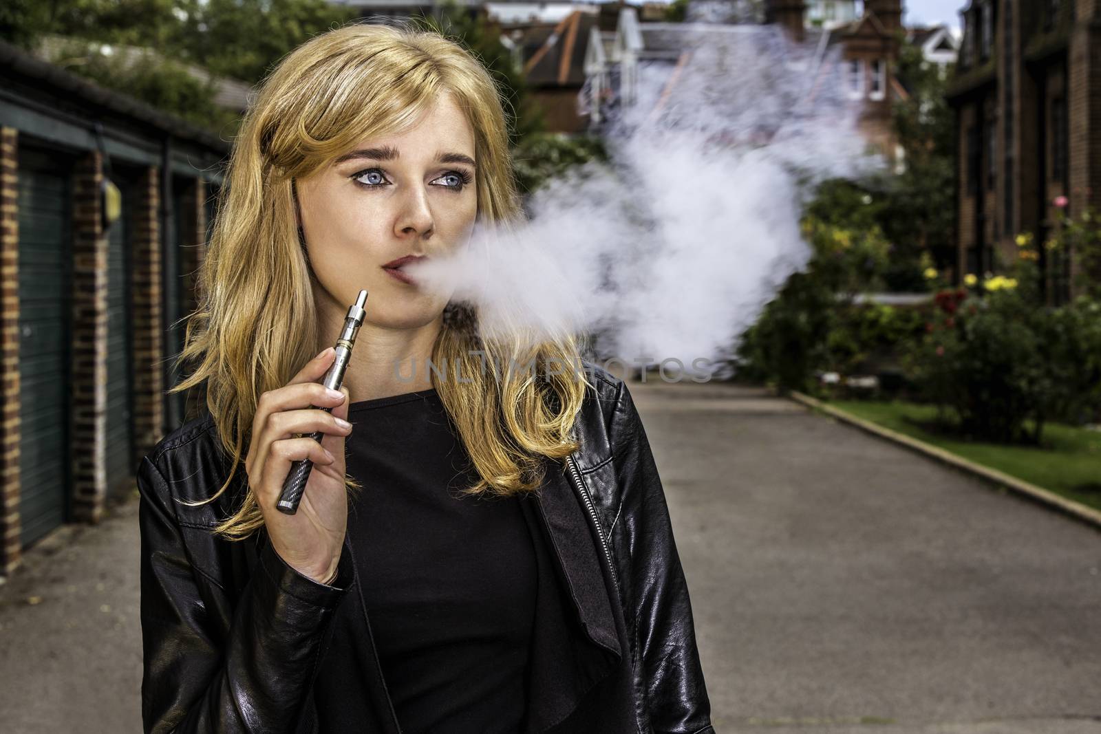 Pretty blond woman smoking an e-cigarette standing in a street in a leather jacket exhaling a cloud a smoke from her mouth while looking off to the side