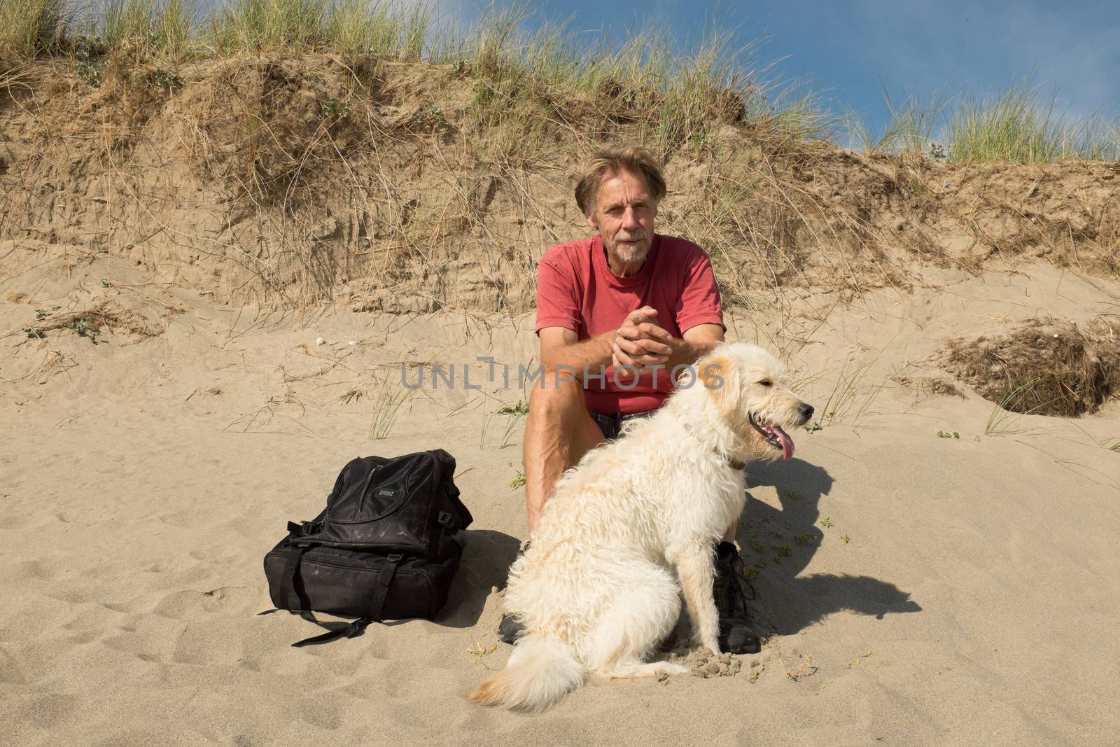 A man and dog, labradoodle, sitting on a beach under a sand dune staring ahead.