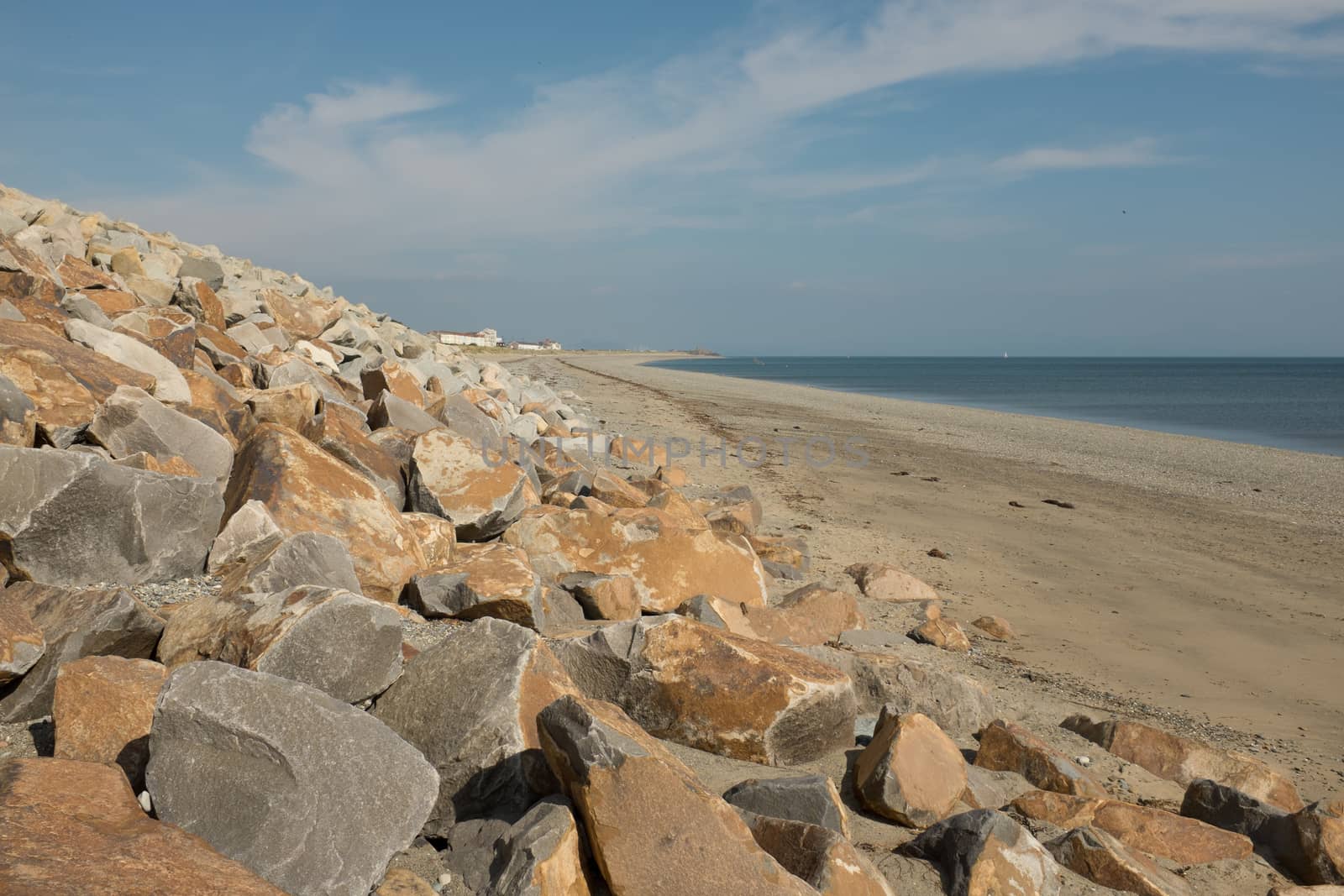 A wall of rocks act as the sea defence on a sandy beach with the sea and blue sky in the distance.