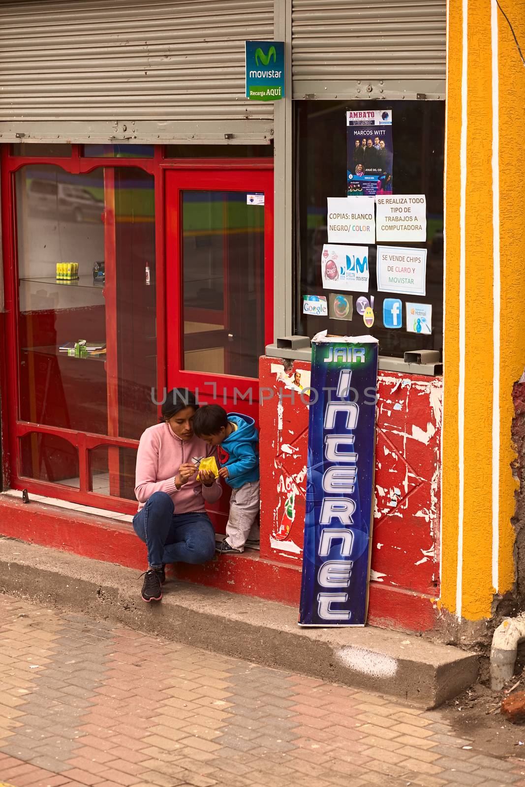 TUNGURAHUA, ECUADOR - MAY 12, 2014: Unidentified woman with child sitting on the stairs at the entrance of an internet cafe and eating a snack in a small town on the road between Ambato and Banos on May 12, 2014 in Tungurahua Province, Ecuador