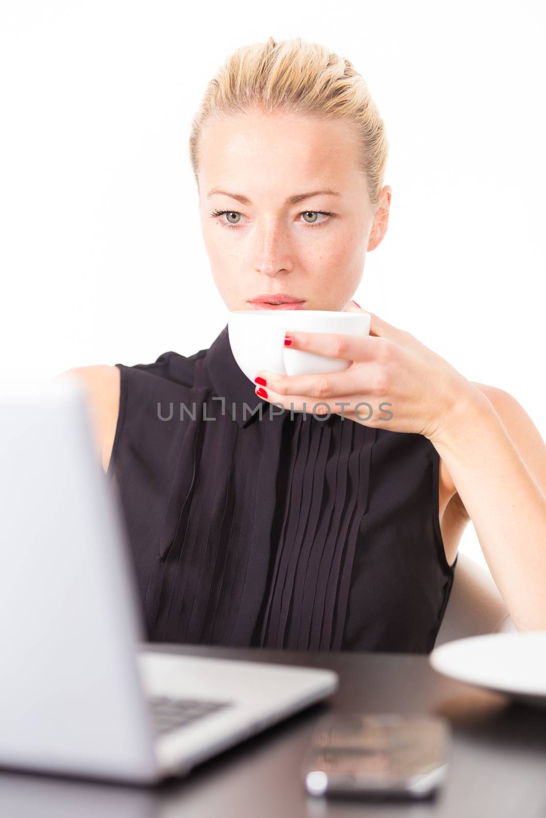 Business woman holding white cup of coffee in her hand while checking some information on her laptop computer in office. Multitasking.