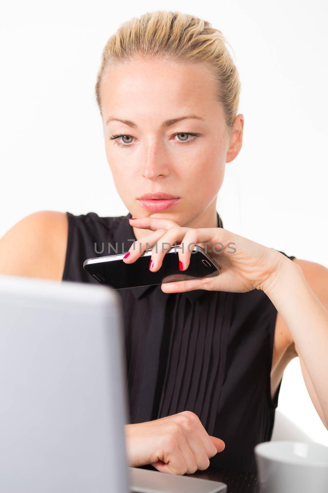 Business woman holding smartphone in her hand, checking some information on her laptop computer in office. 