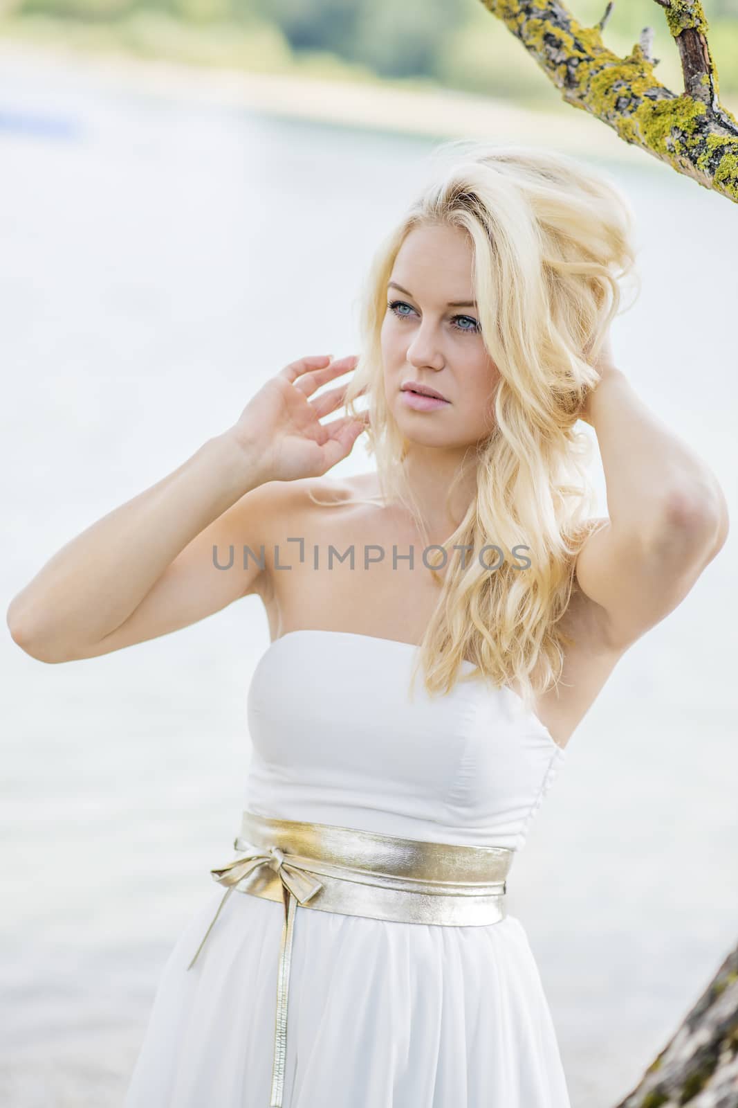 Blond young woman standing under a tree at the edge of a lake