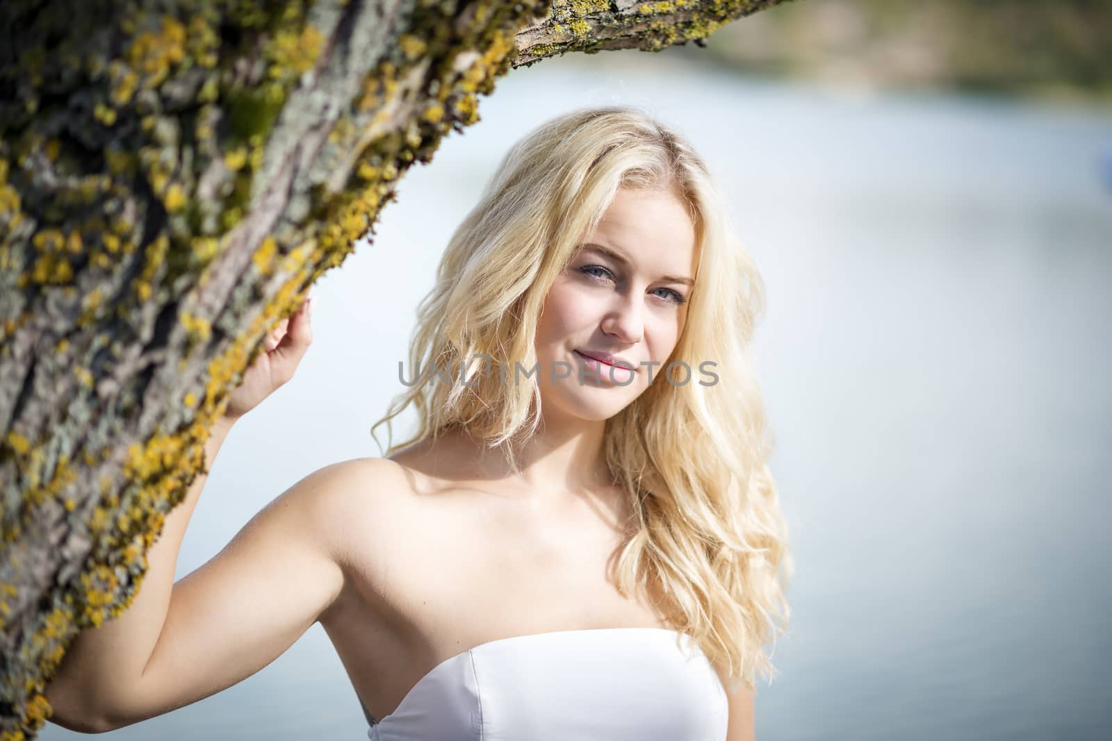 Blond young woman standing under a tree at the edge of a lake