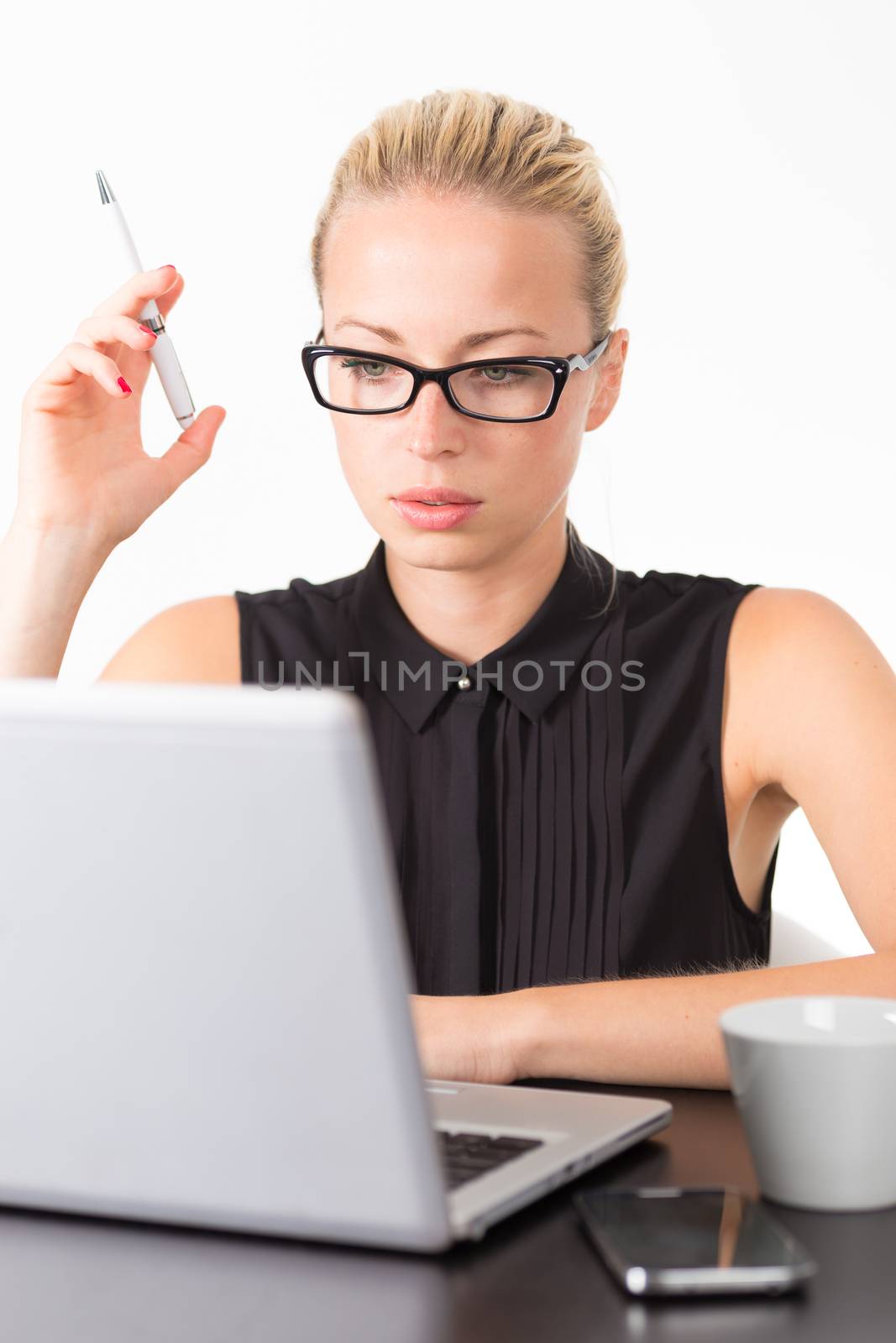 Business woman wearing black glasses, holding a pen in her hand, checking some information on her laptop computer in office. 