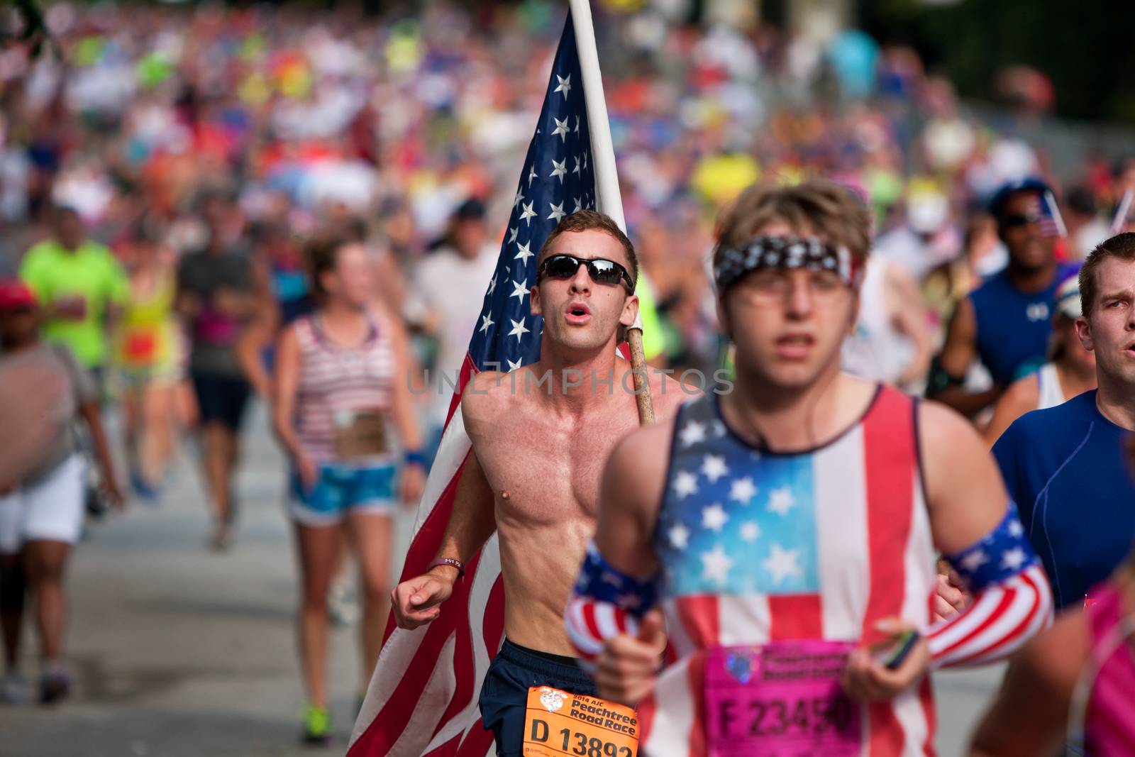 Atlanta, GA, USA - July 4, 2014:  A young man carries a large American flag on his shoulder as he runs among thousands of other people nearing the finish line of the Peachtree Road Race.