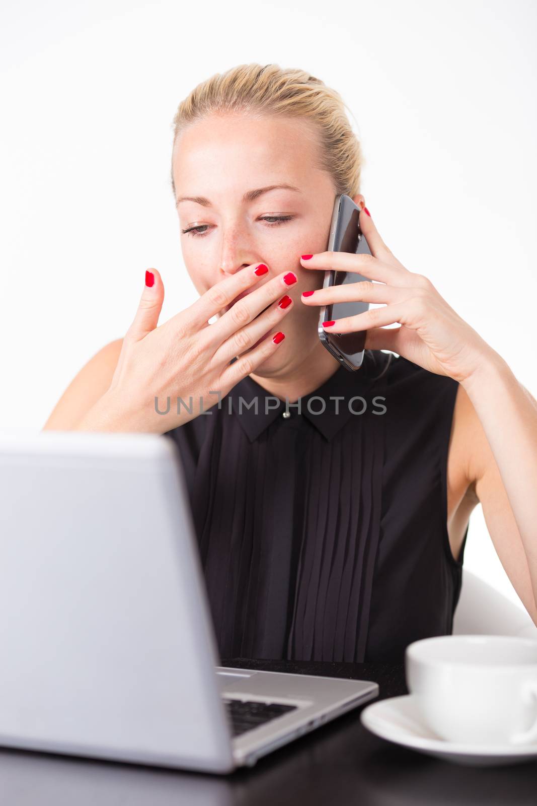 Work late into the night. Business woman yawning while talking on the mobile home and checking some information on her laptop computer in office. White cup of coffee on the desk. 