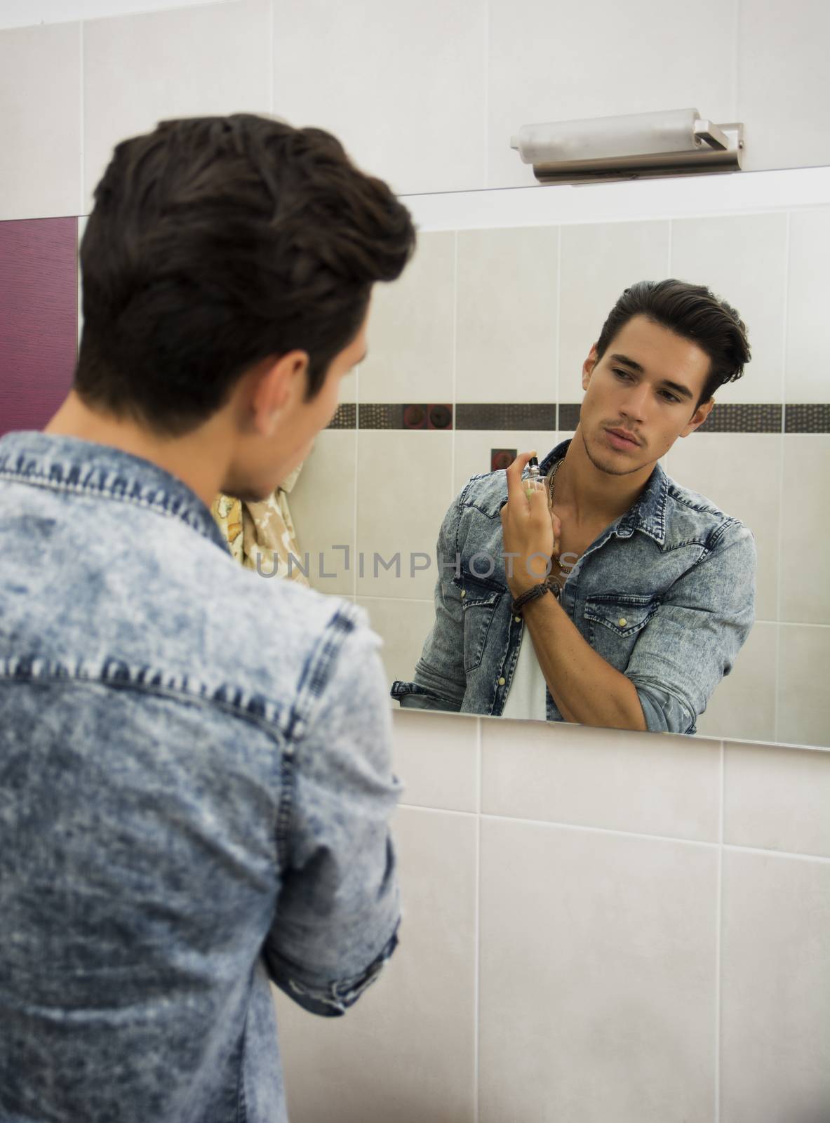 Attractive young man looking at bathroom mirror, spraying cologne or perfume