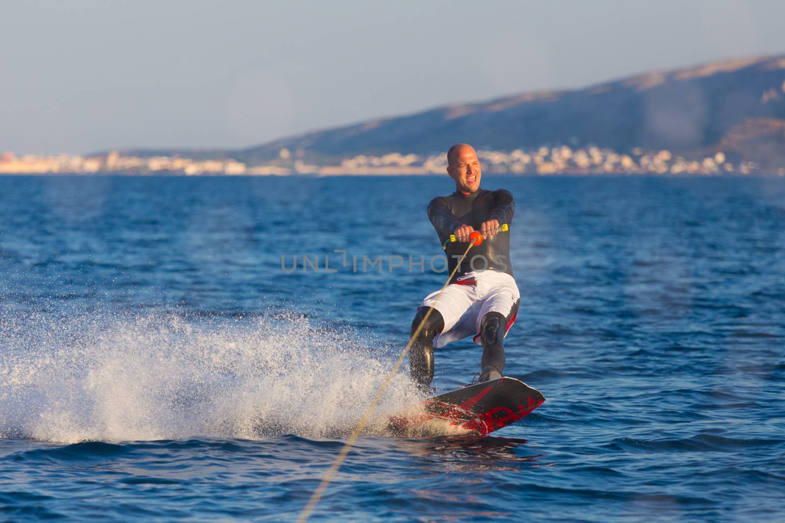 Wakeboarder in wetsuit riding in sunset. Wakeboarding is a surface water sport which involves riding a wakeboard over the surface of a body of water.