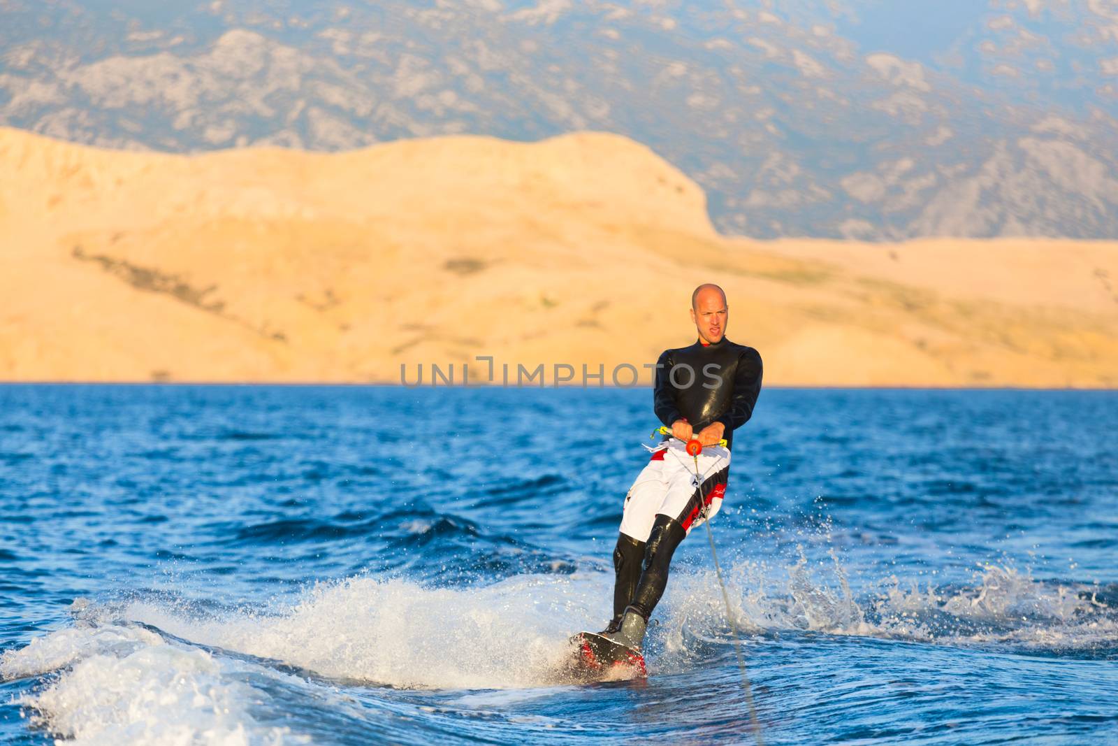 Wakeboarder in wetsuit riding in sunset. Wakeboarding is a surface water sport which involves riding a wakeboard over the surface of a body of water.