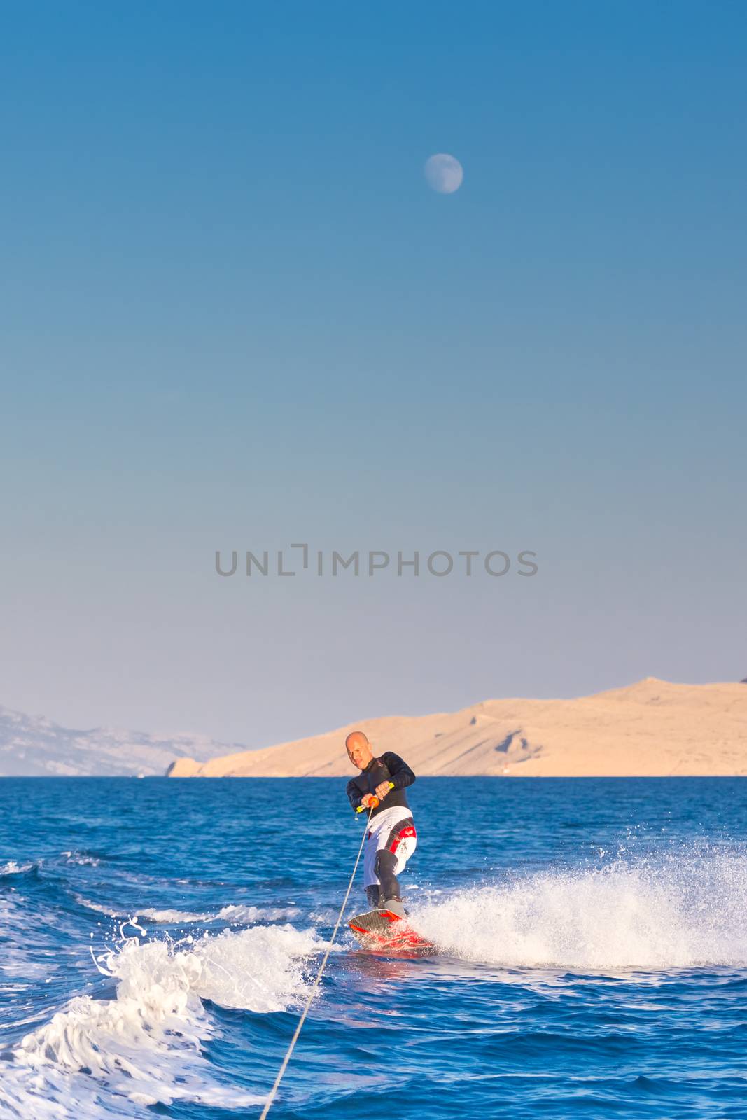 Wakeboarder in wetsuit riding in sunset. Wakeboarding is a surface water sport which involves riding a wakeboard over the surface of a body of water.