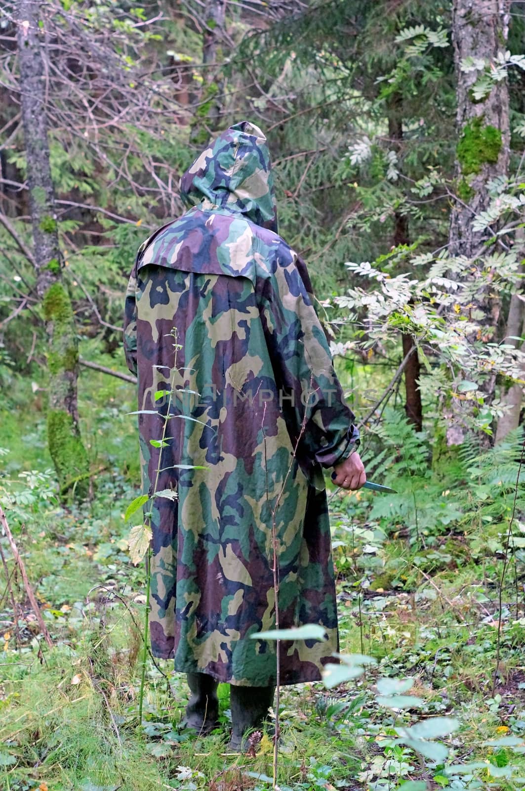 A man in a camouflage hooded standing back in the woods with a knife in his hand               