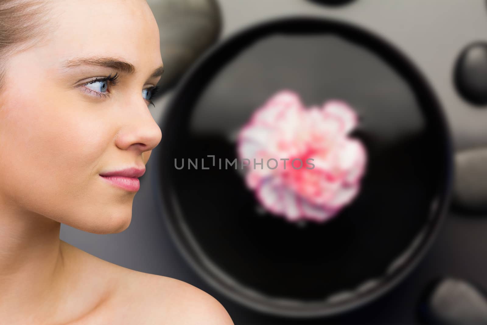 Smiling blonde natural beauty against pink flower floating in bowl with pebbles