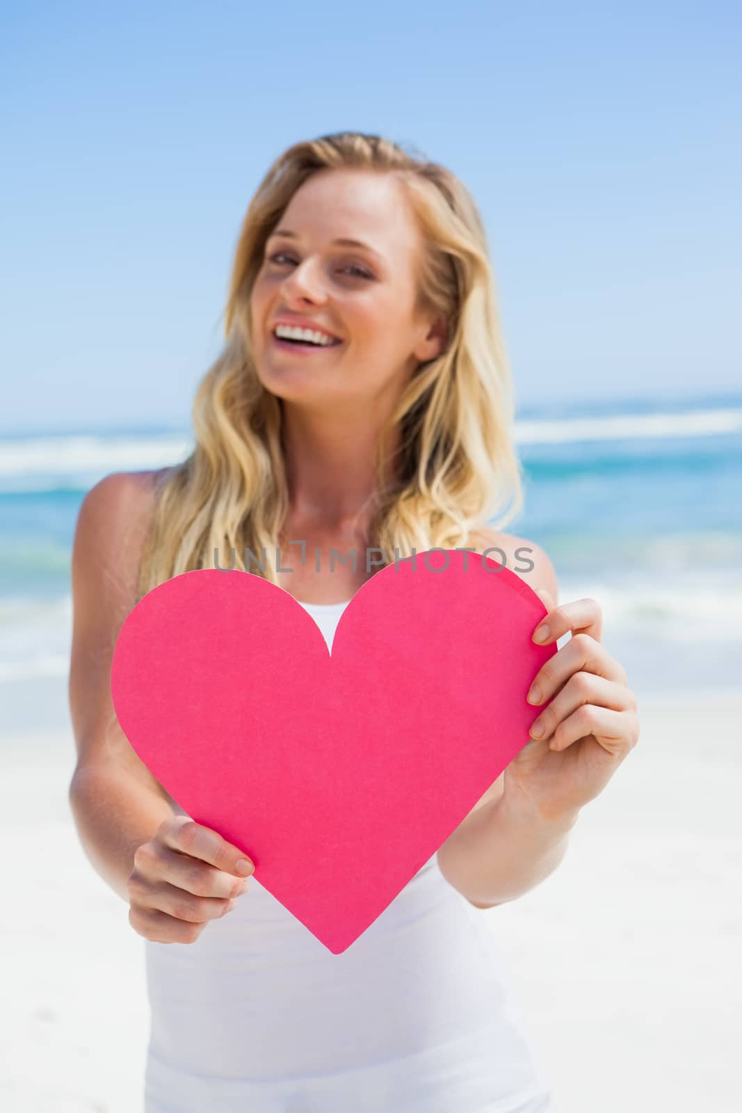 Smiling blonde showing pink heart on the beach on a sunny day