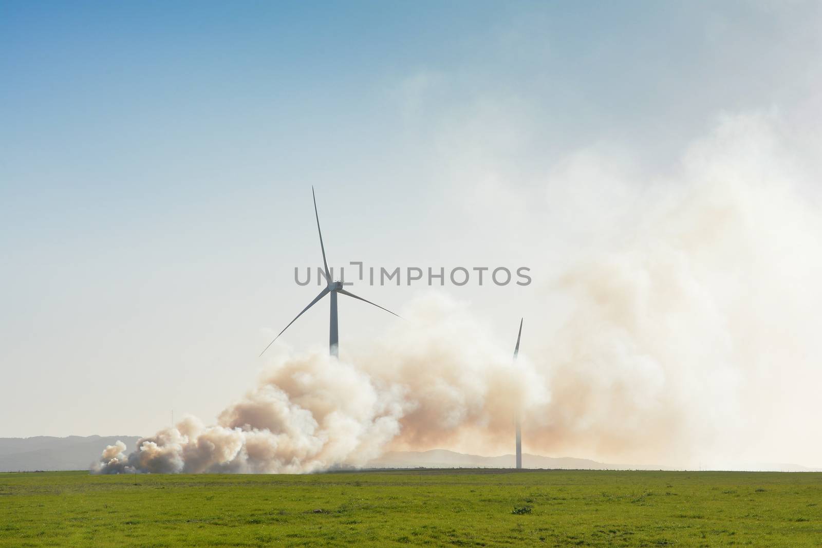 A small wind farm in a green field with a controlled fire burning close by. Thick clouds of smoke conceal most of the nearest turbines.