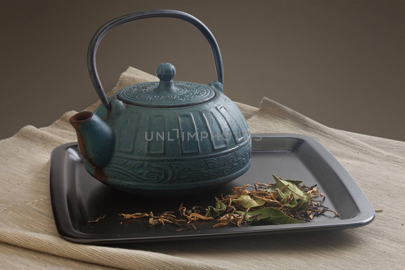 Black plate with blue teapot and green dried tea on the napkin