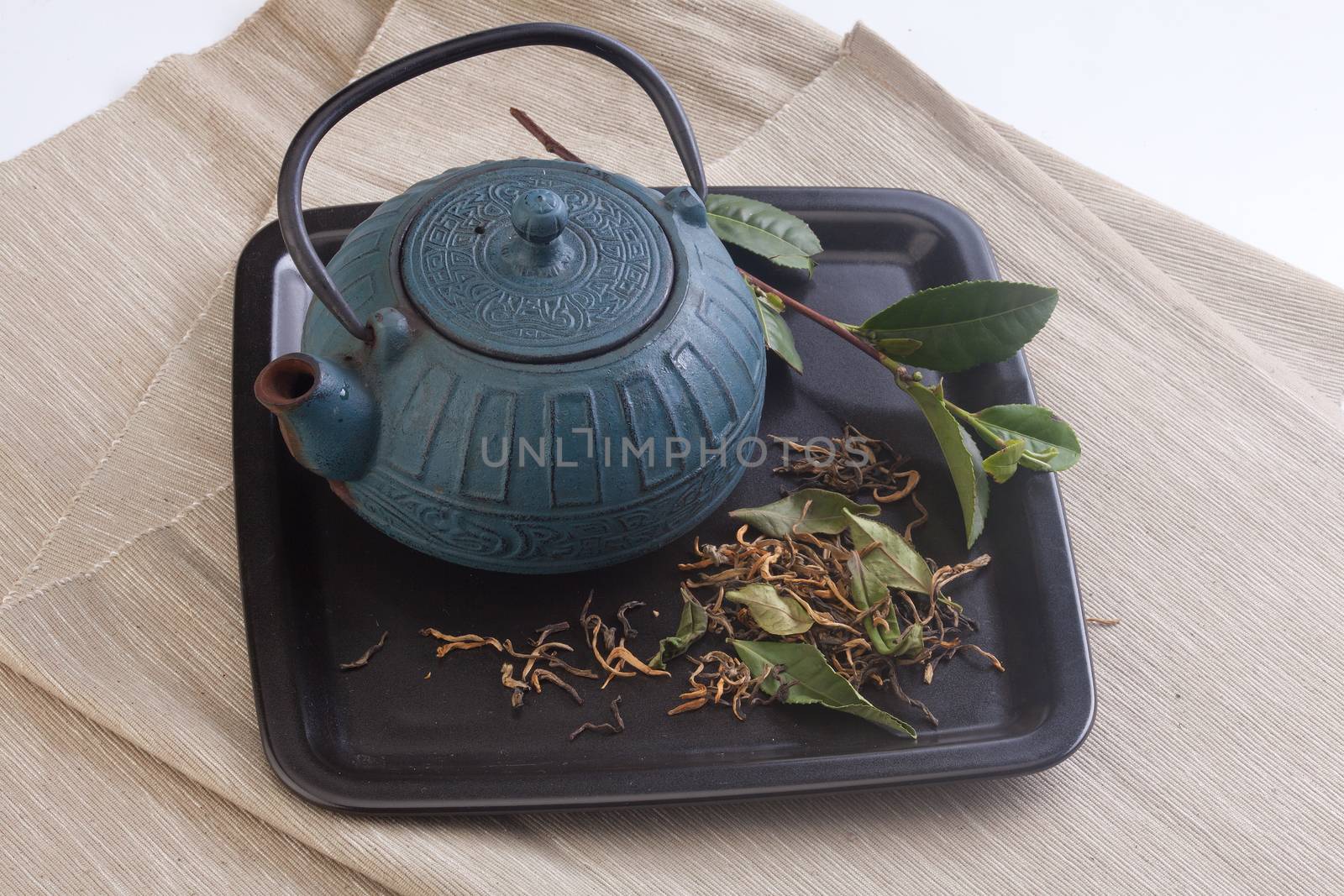 Black plate with blue teapot, green tea branch and dried tea leaves on the napkin