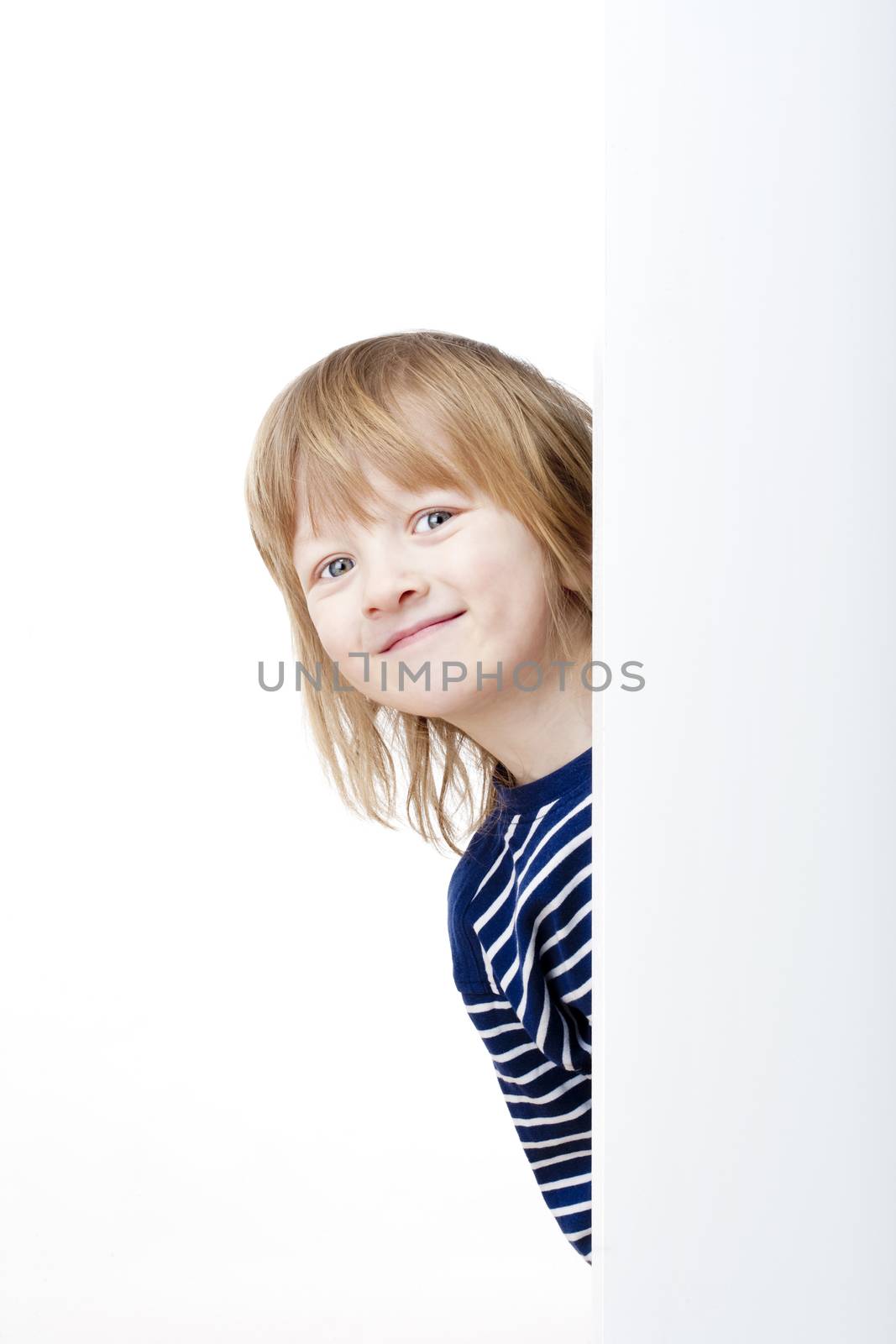 curious boy with long blond hair peeking out from behind a white board smiling