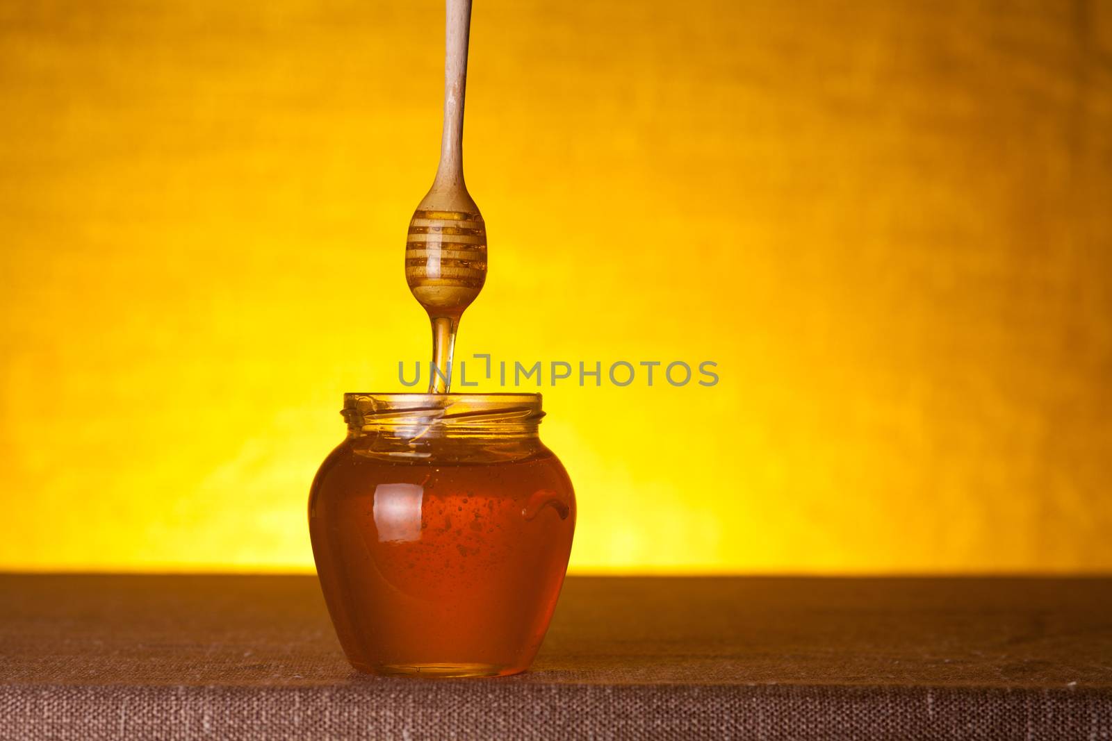 Honey jar with wooden dipper and flowing honey, studio shot over yellow background 