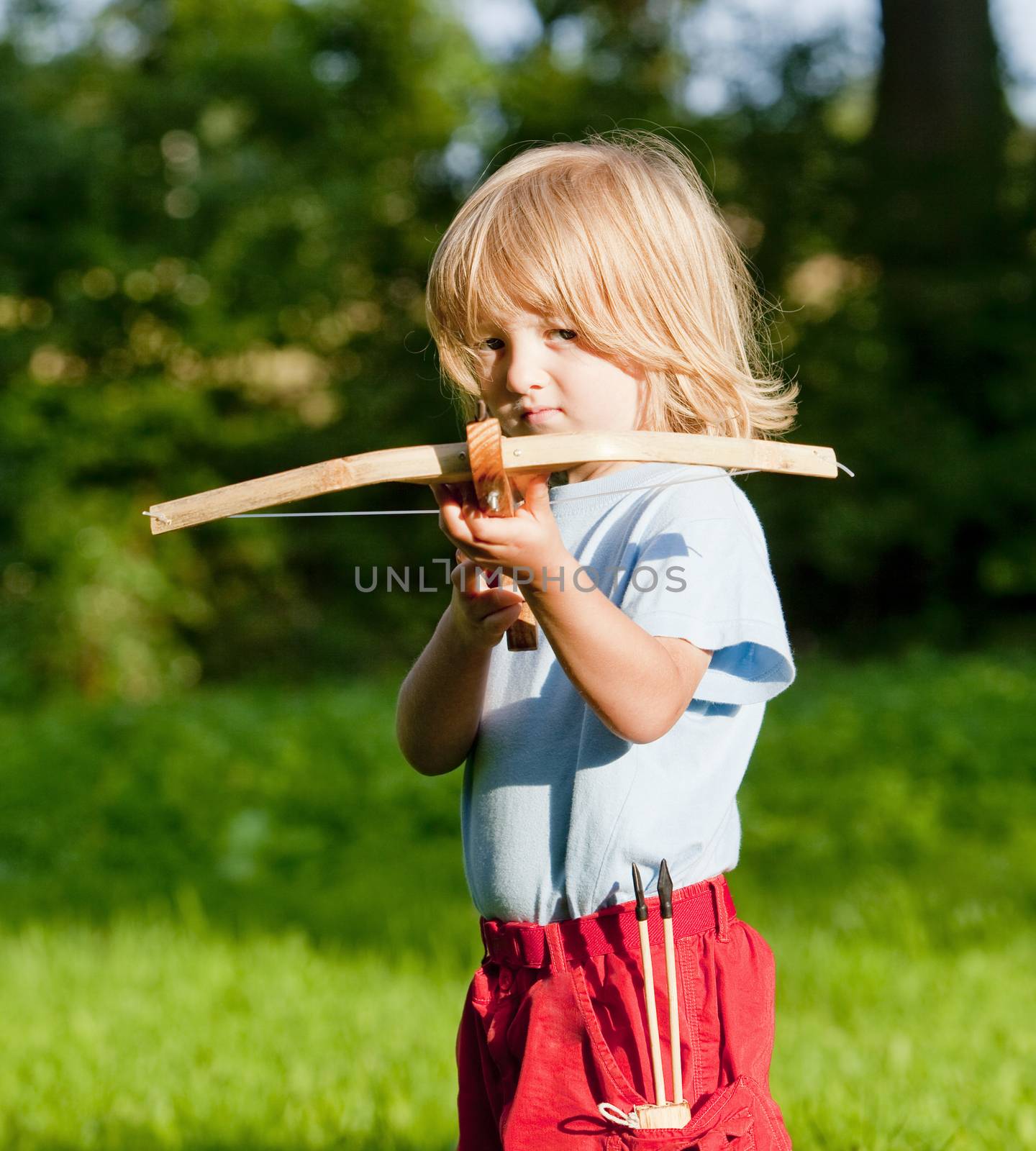 little boy with long blond hair shooting with toy crossbow in the garden