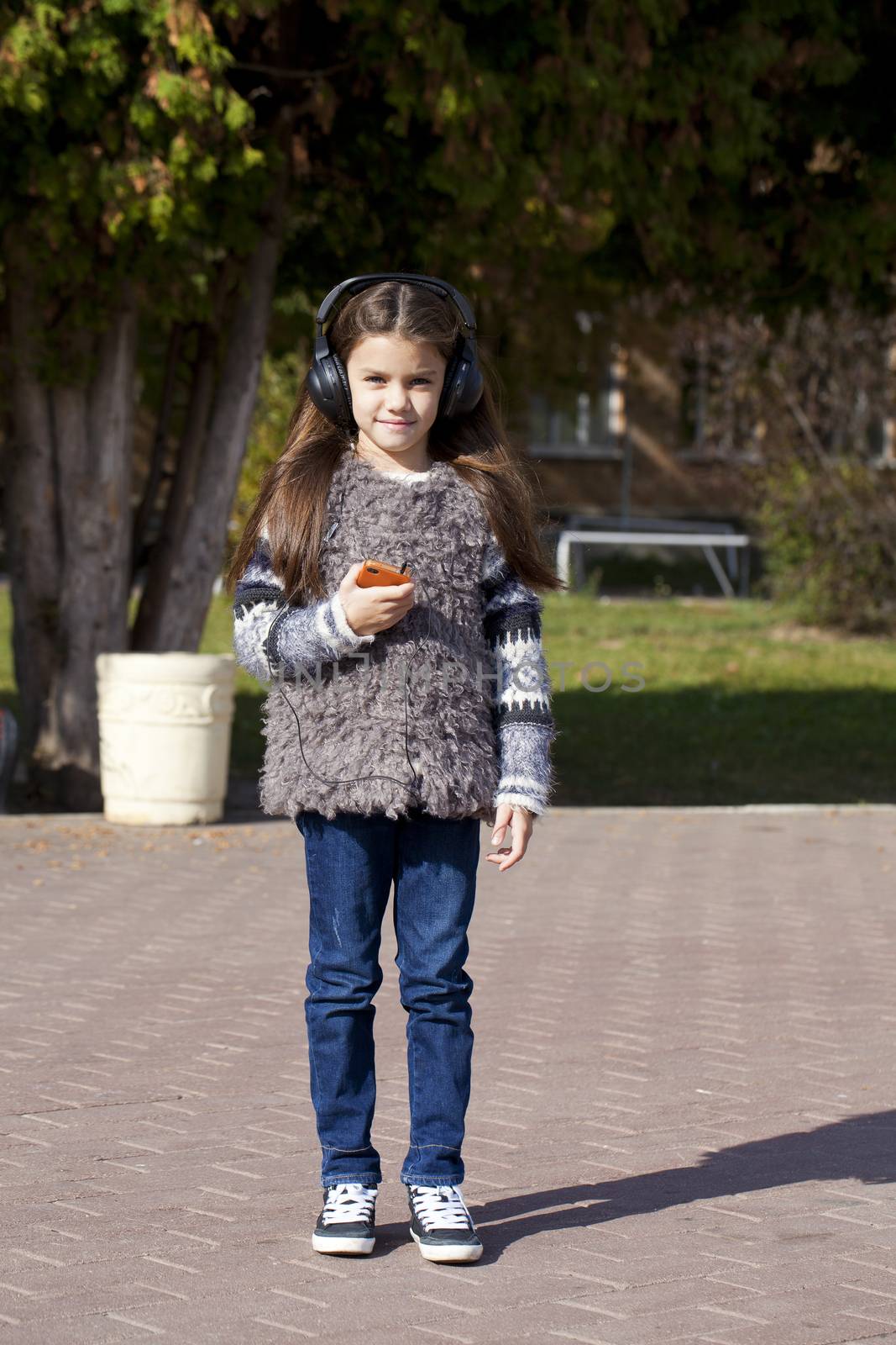 Beautiful little girl listening to music on headphones in autumn park