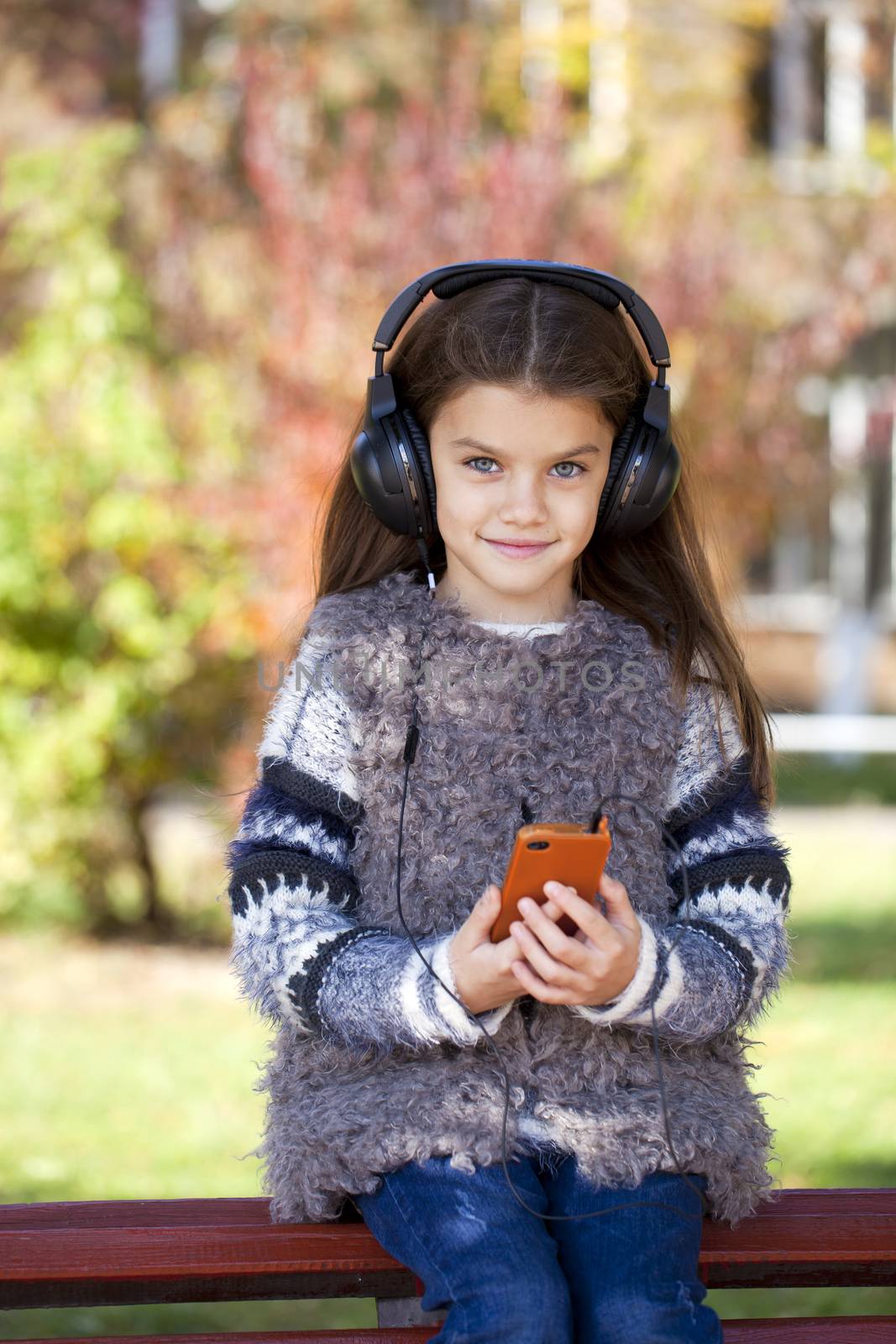 Beautiful little girl listening to music on headphones in autumn park