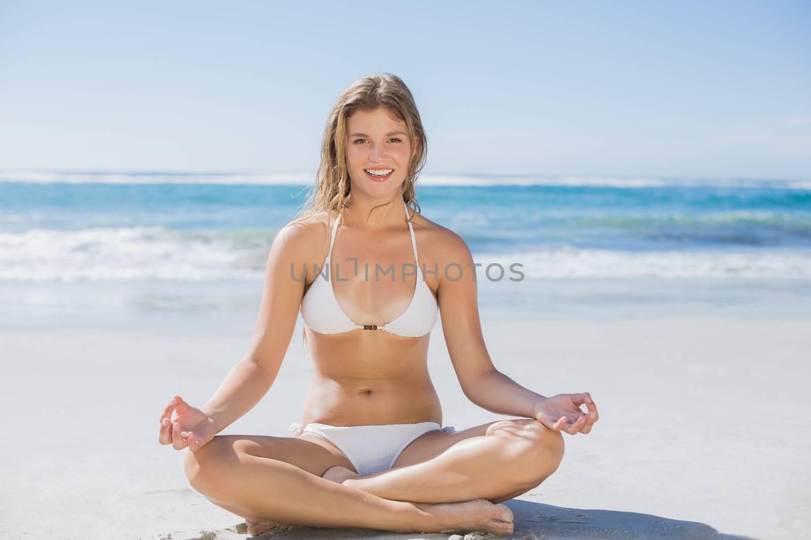 Beautiful girl in white bikini sitting in lotus pose on beach on a sunny day