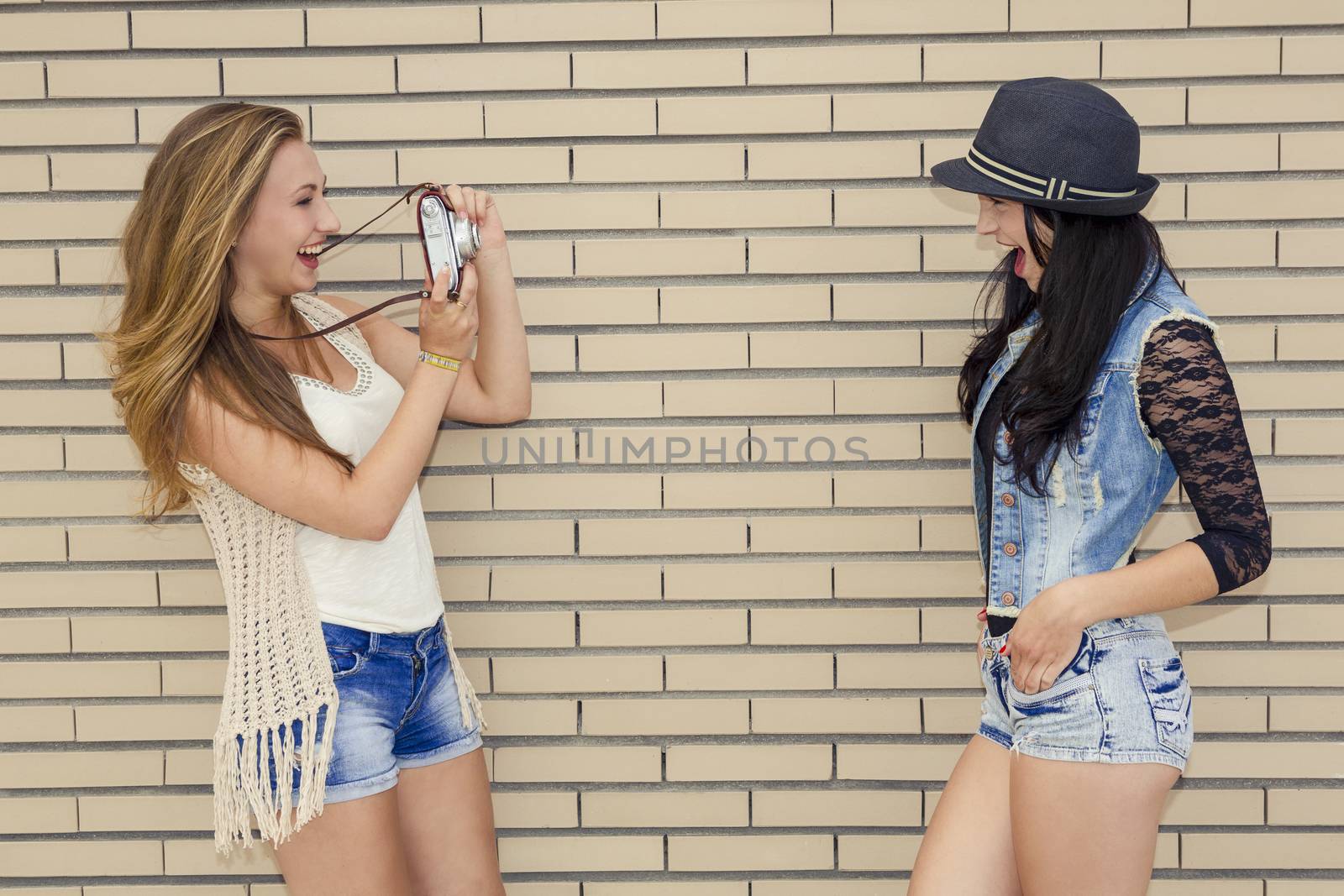 Two beautiful and young girlfriends taking pictures, in front of a brick wall