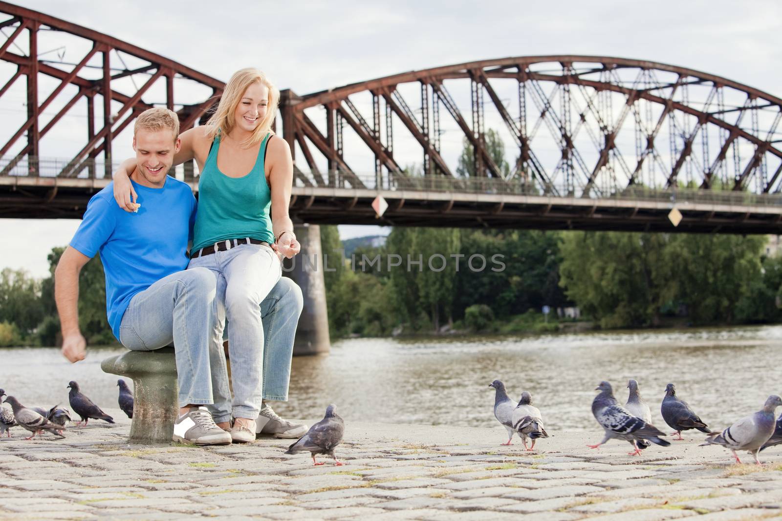 young happy couple sitting by the riverside, feeding birds