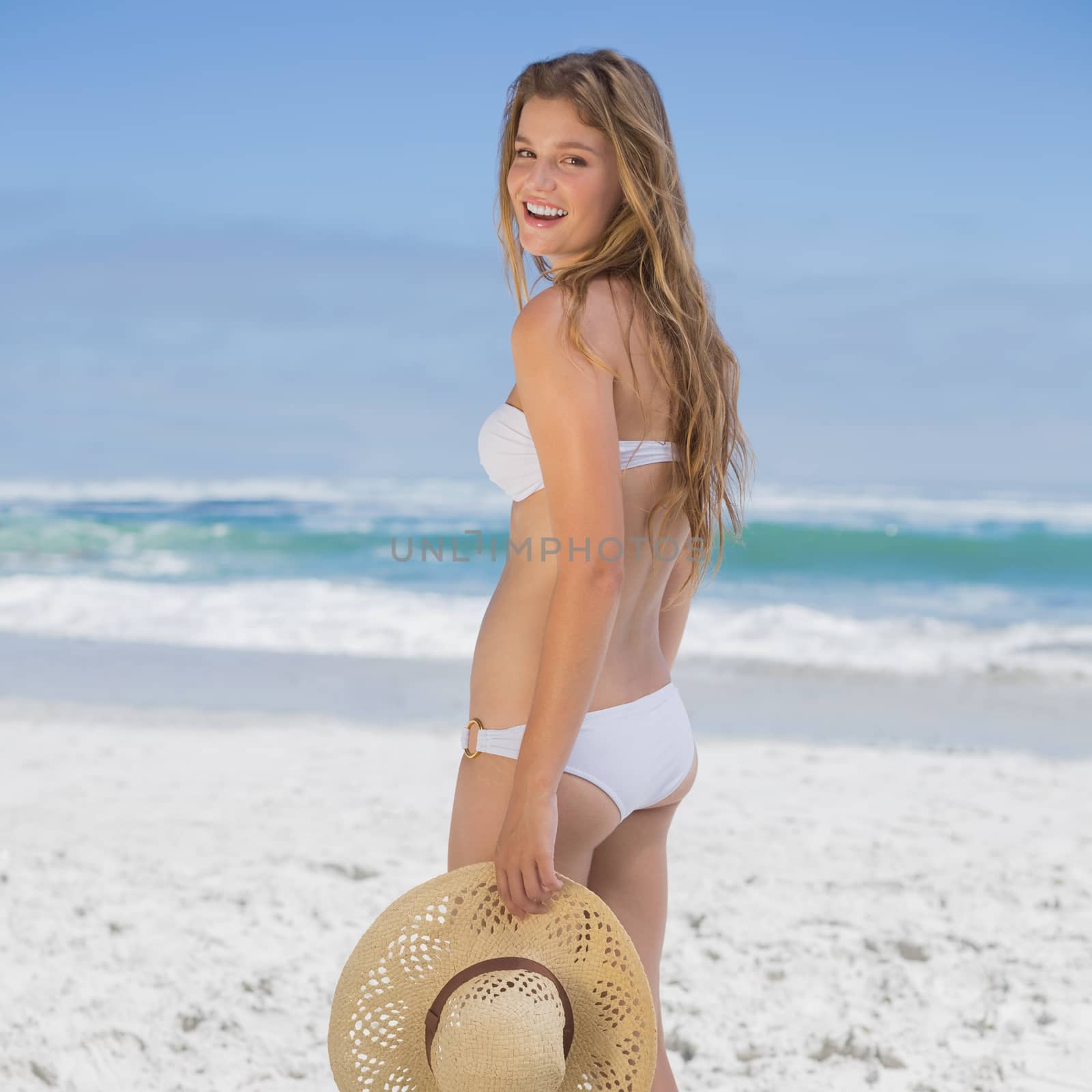Beautiful happy blonde on the beach in white bikini holding sunhat on a sunny day