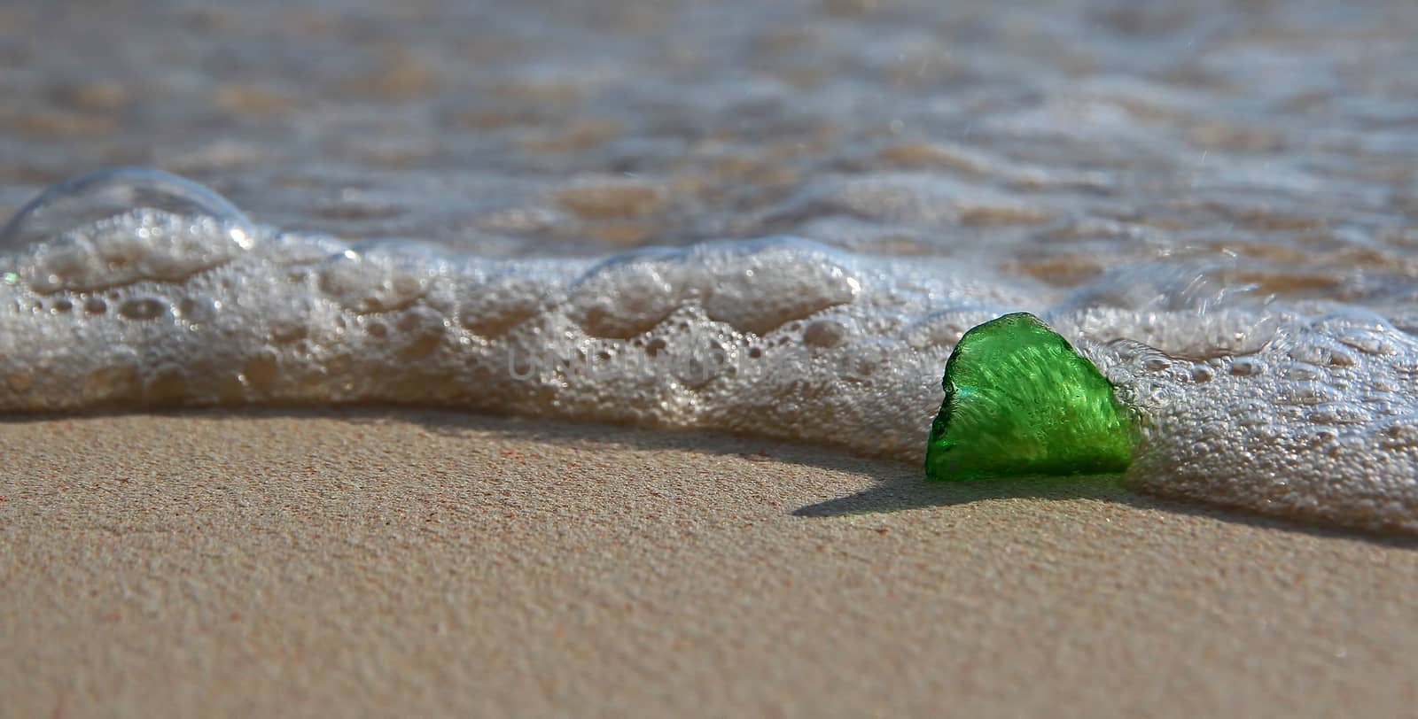 A close up of a heart shaped, single piece of green sea glass, on a tropical beach, with a breaking wave near the glass