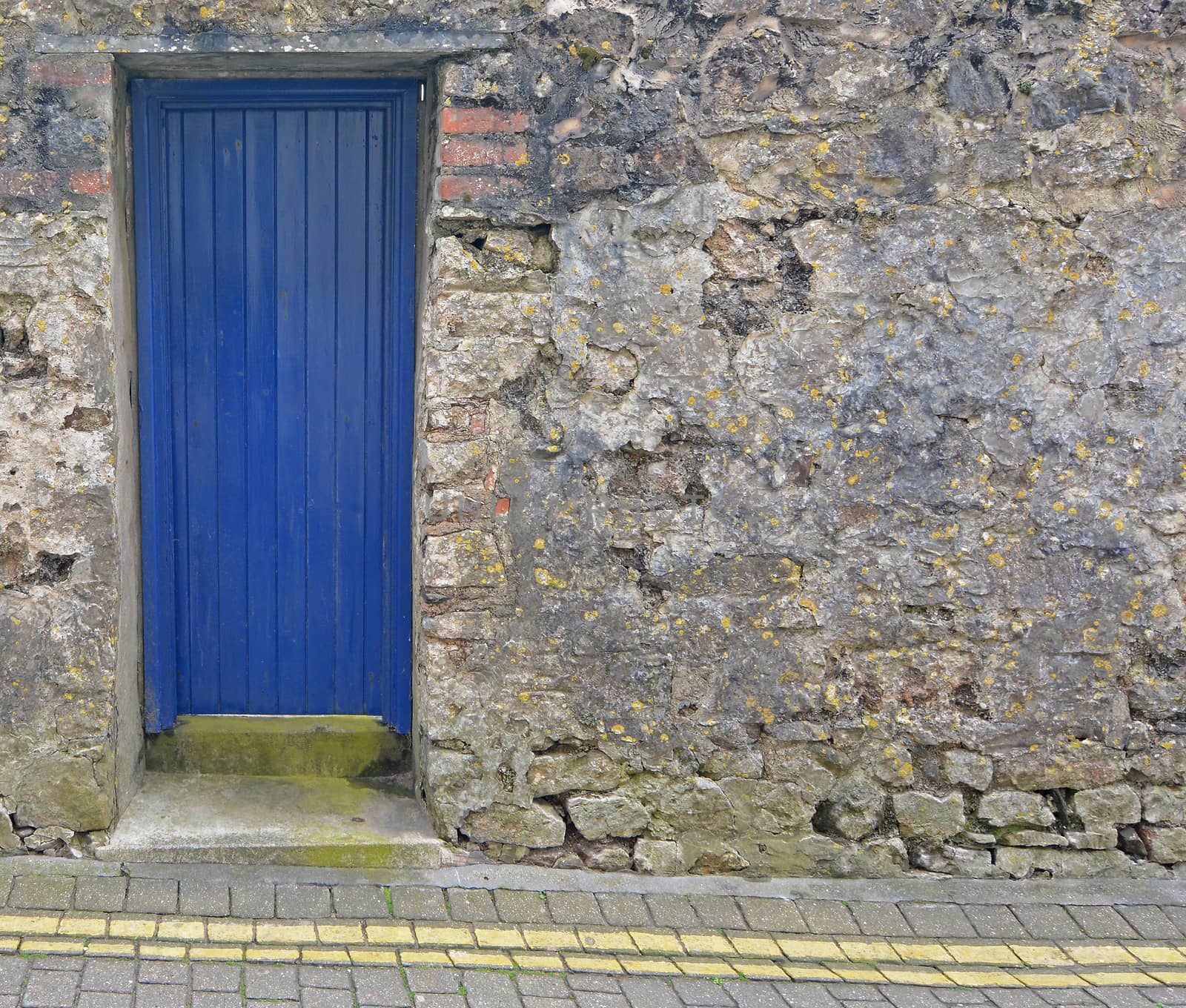 A blue, wooden door, set into a stone wall, in front of double yellow lines