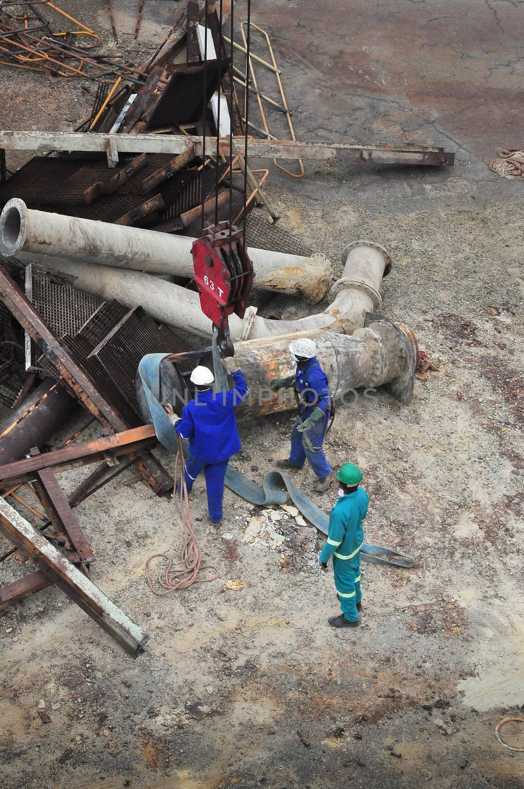Workers on the ground, seen from above, busy either hooking up or unhooking large structural pieces of equipment with a crane hook overhead.