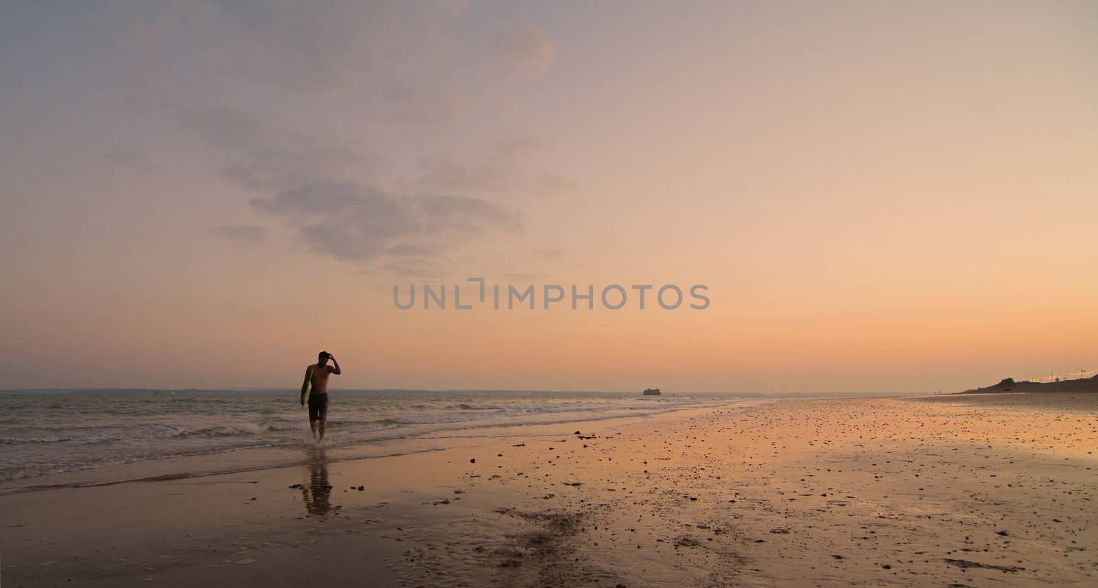 A single surfer walking along the shore, with the sun setting behind him