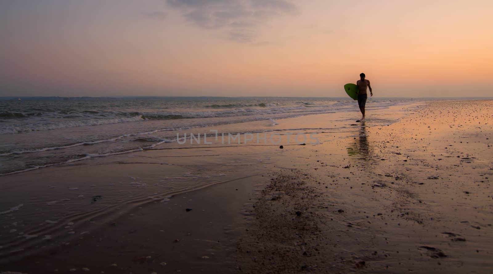 A single surfer walking along the shore, with the sun setting behind him