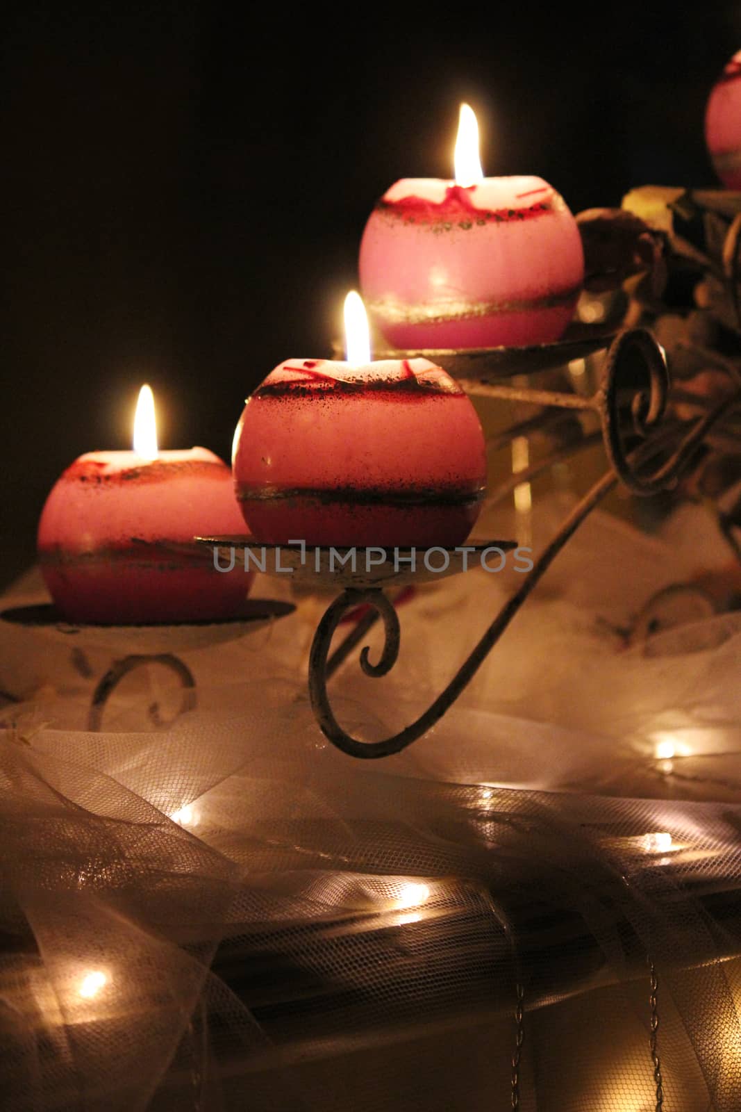 Round pinkish, lit candles on curved decorative stand. Set on a table with small decorative lights underneath the netting.