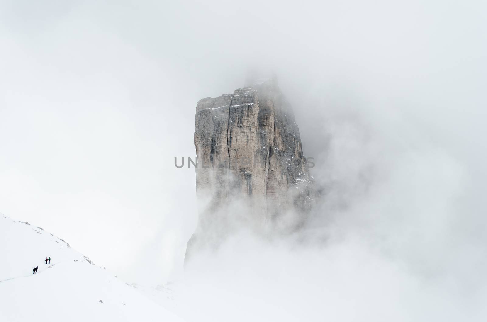 4 people in the lower left corner hiking through the snow in winter around the Tre Cime di Lavaredo, Drei Zinnen, Dolomites mountain peaks partially covered in the clouds.