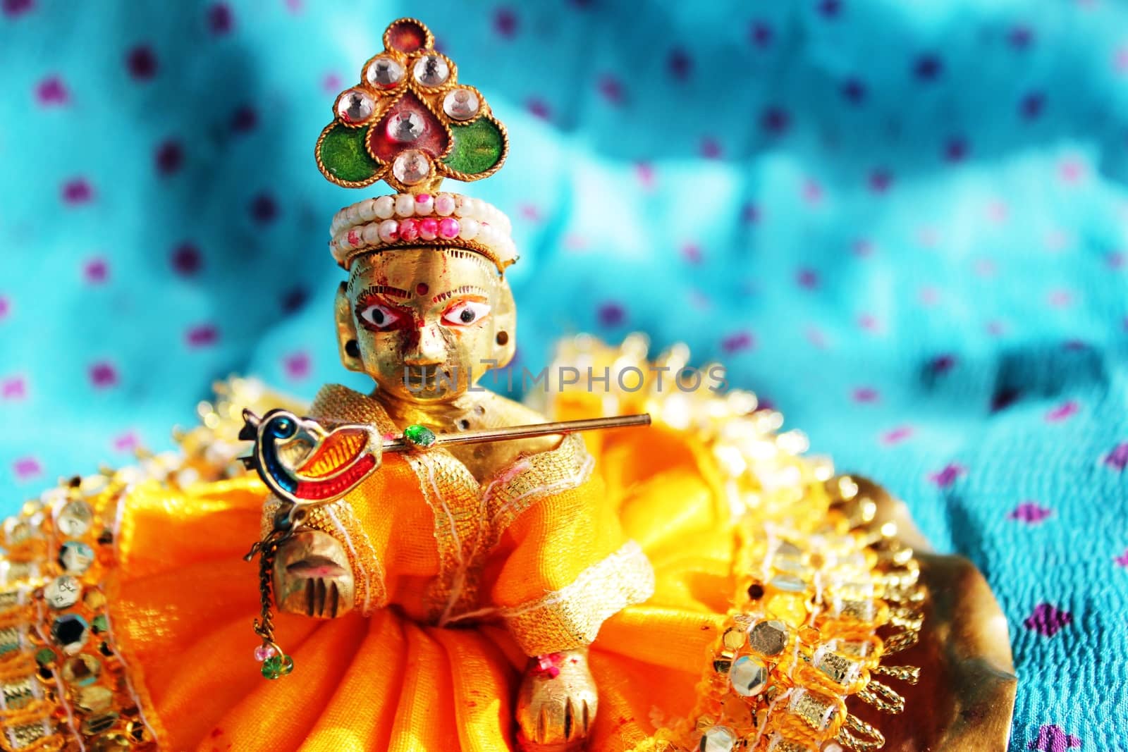 Brass idol of baby lord Krishna, a Hindu god, dressed in bright yellow clothing, against a polka dotted aqua colored cloth background, sporting his legendary musical flute. Ideal for Hindu religious use and festivals - like Janamashtami, Gokul ashtami. 

