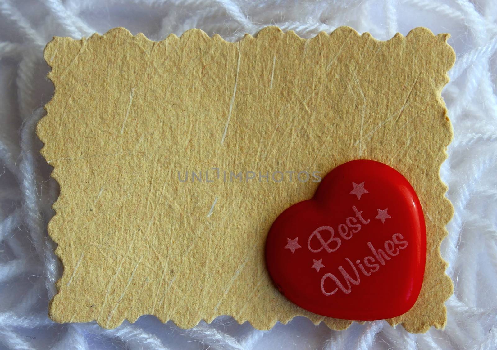 A blank hand made paper message note card, with a tiny red plastic heart that reads ' best wishes', against strands of white wool yarn
