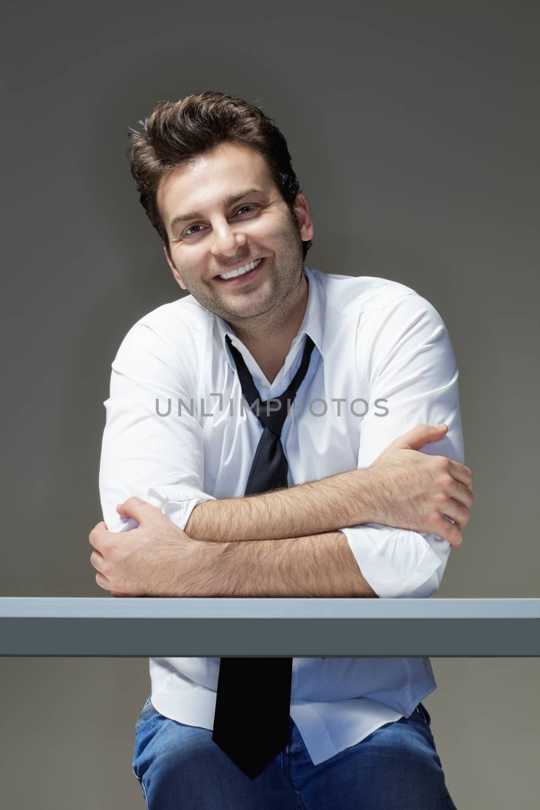 businessman in shirt, tie and jeans sitting at desk, smiling - isolated on gray