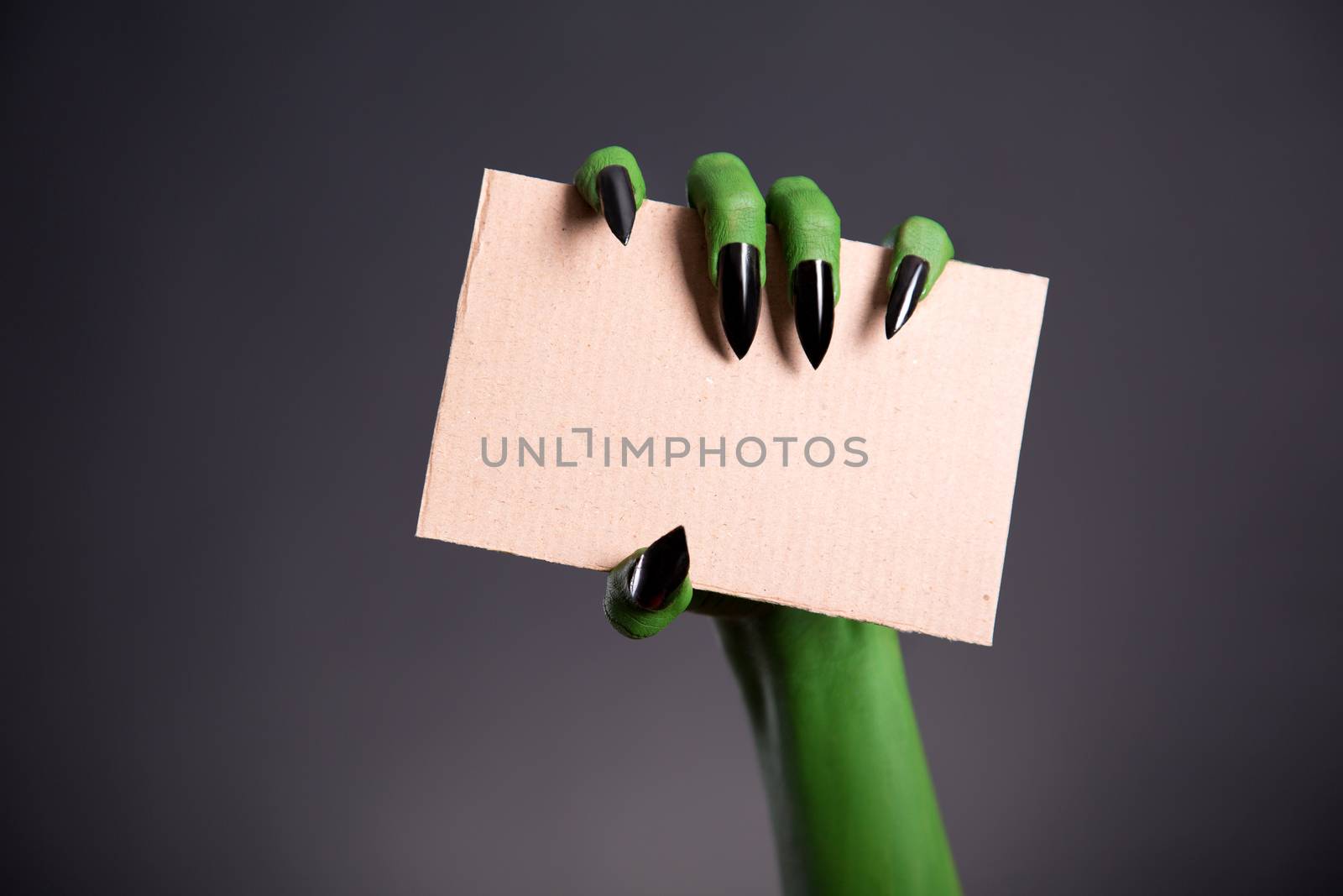 Green monster hand with sharp nails holding blank piece of cardboard, Halloween theme  