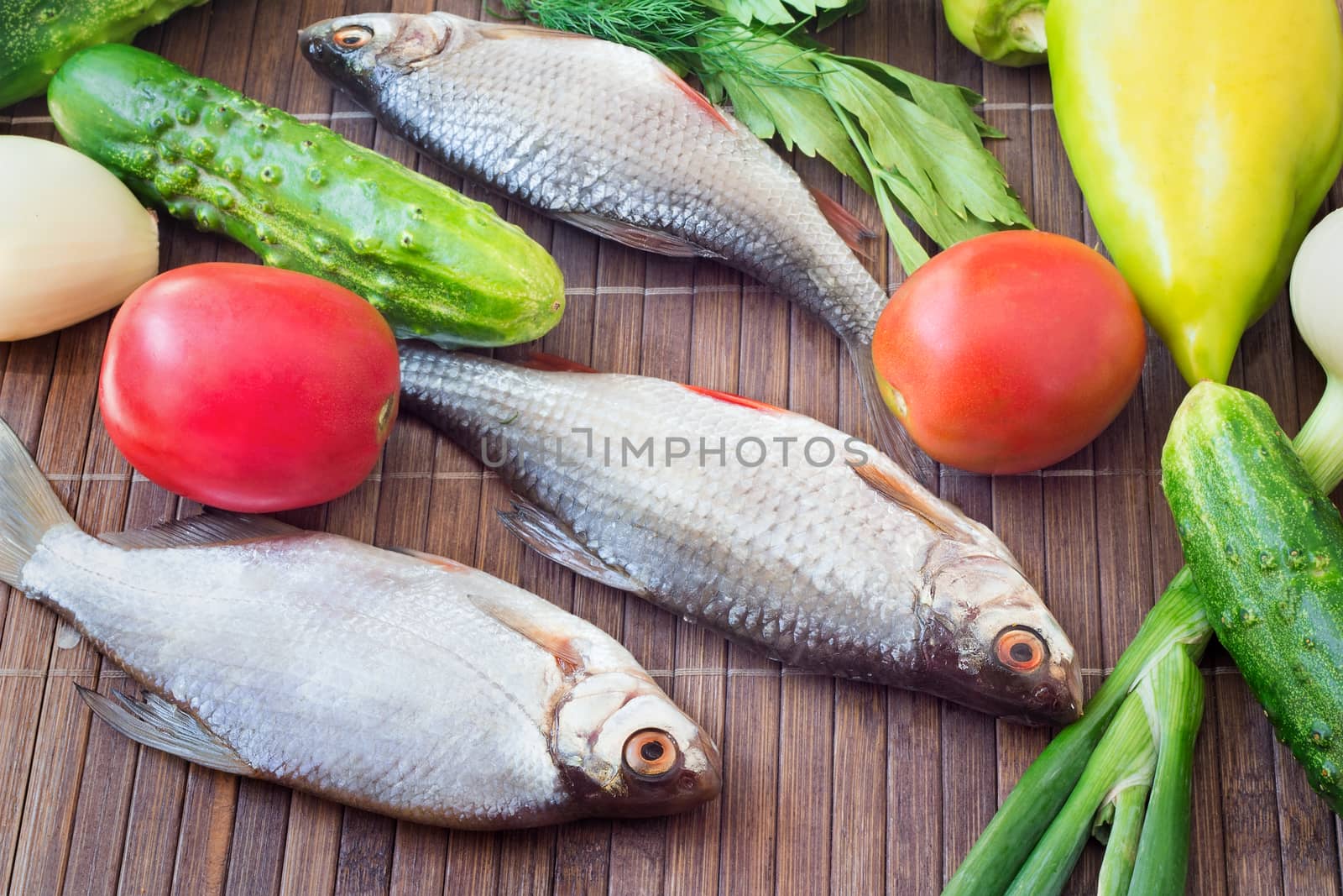 On a table on a chopping board there are a fish, cucumbers, tomatoes, onions, spices and parsley greens.